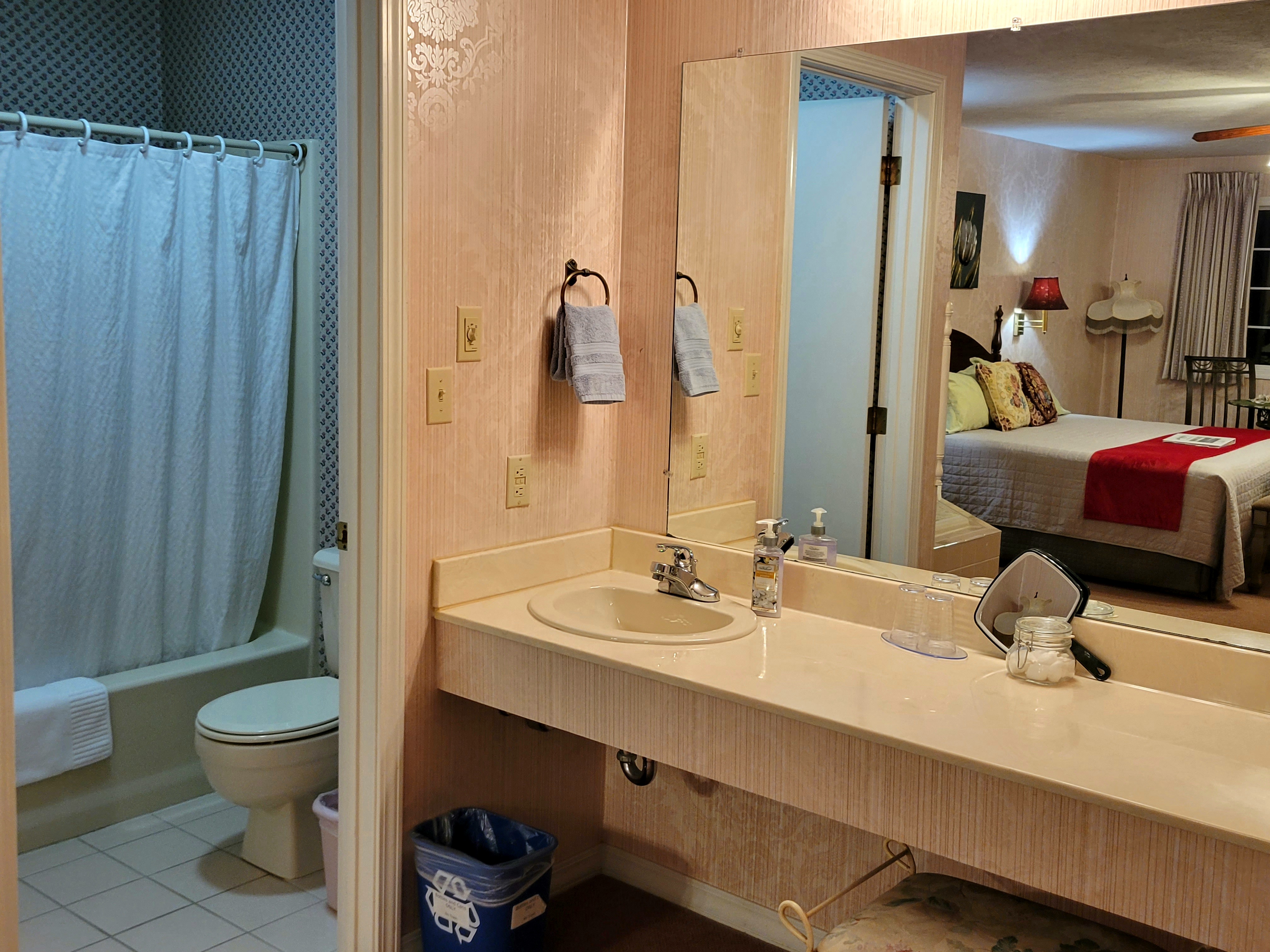 View looking into the bathroom space that features a toilet and a shower curtain where the shower/tub combo is in the room. The water closet is wallpapered and clean. The corner of the vanity right outside the water closet is also visible.