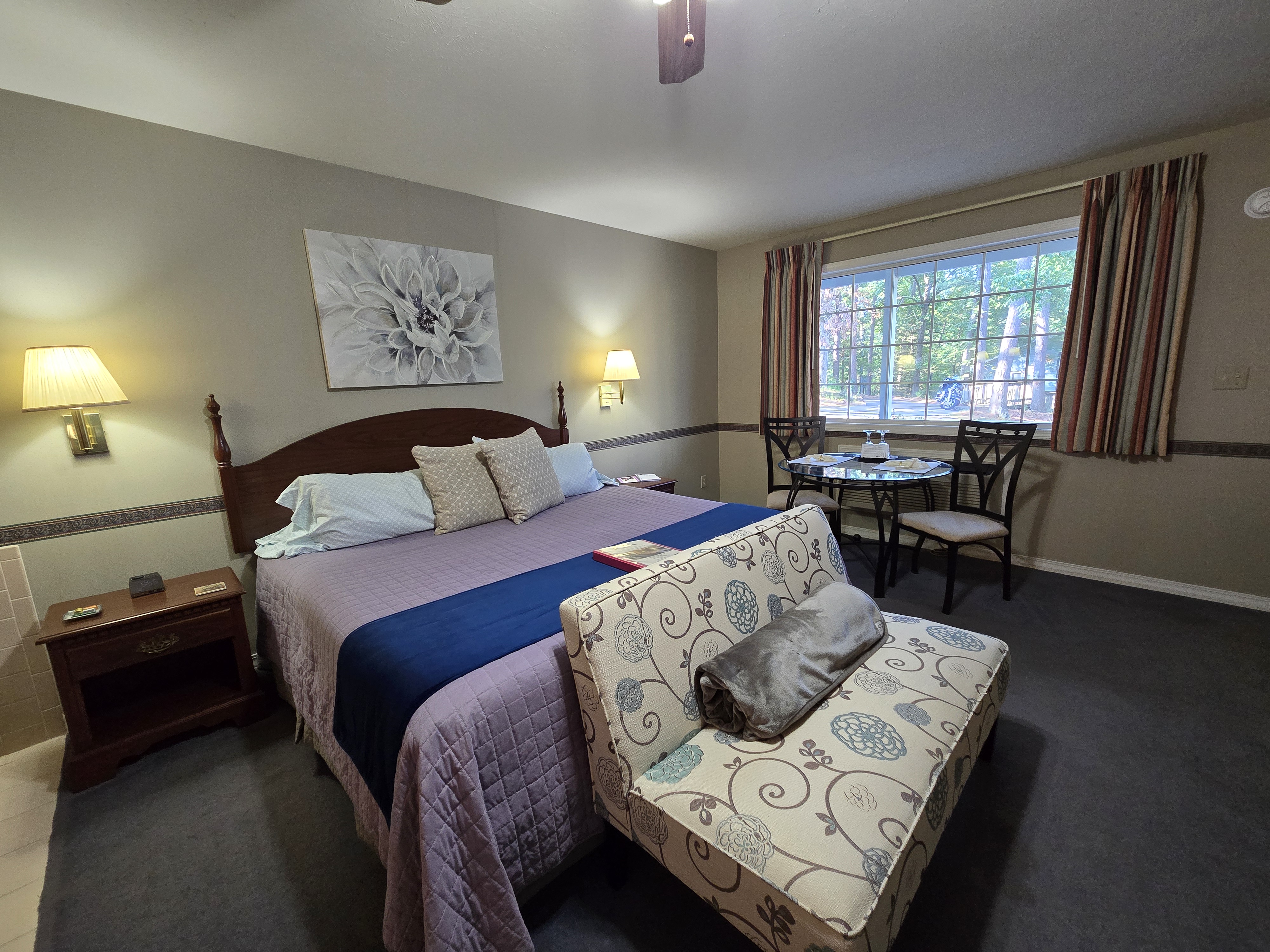 room with a king bed, chaise lounge, small glass dining table, jetted tub in the corner, and a view into the bathroom vanity area.