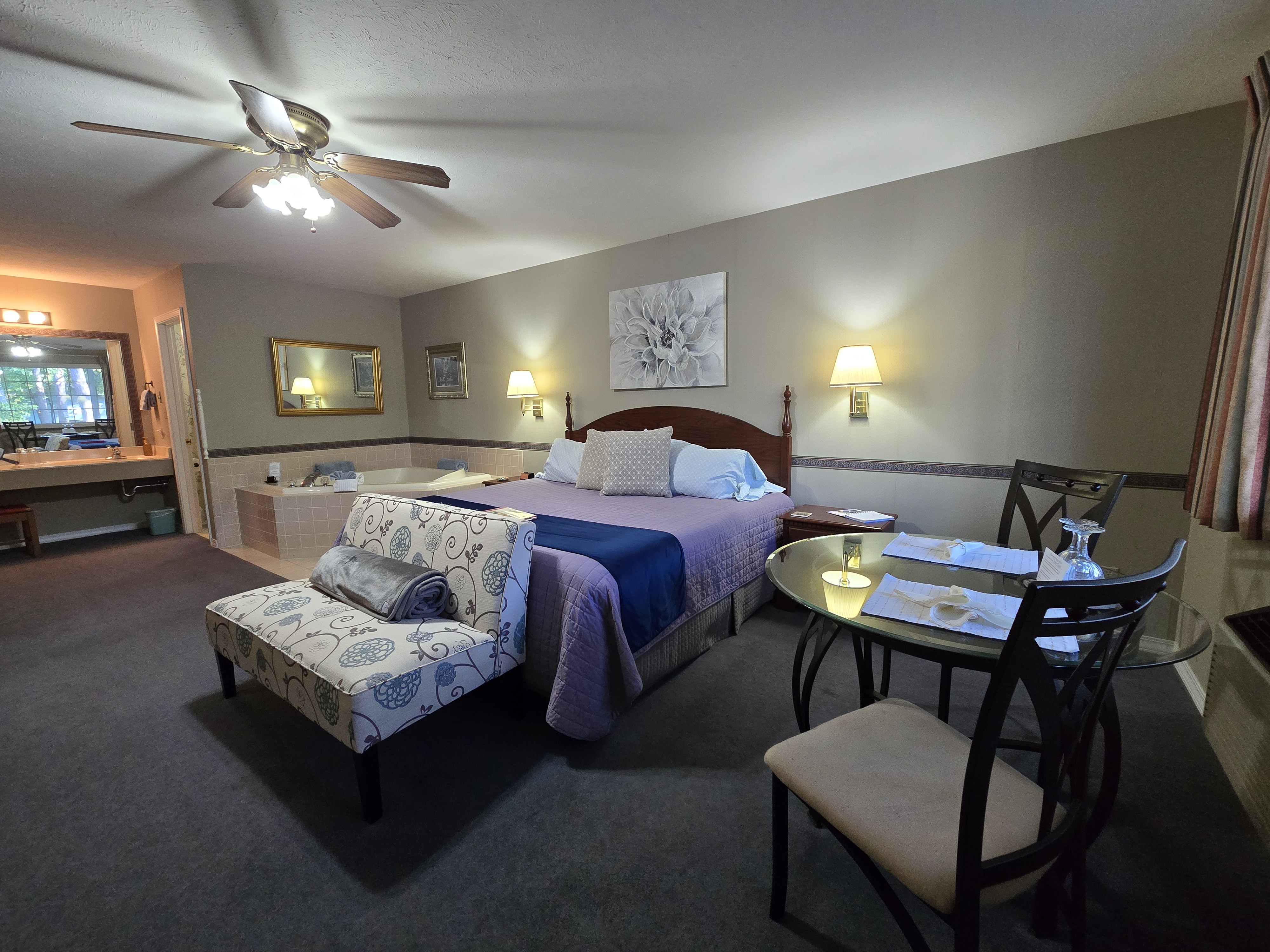 room with a king bed, chaise lounge, small glass dining table, jetted tub in the corner, and a view into the bathroom vanity area.