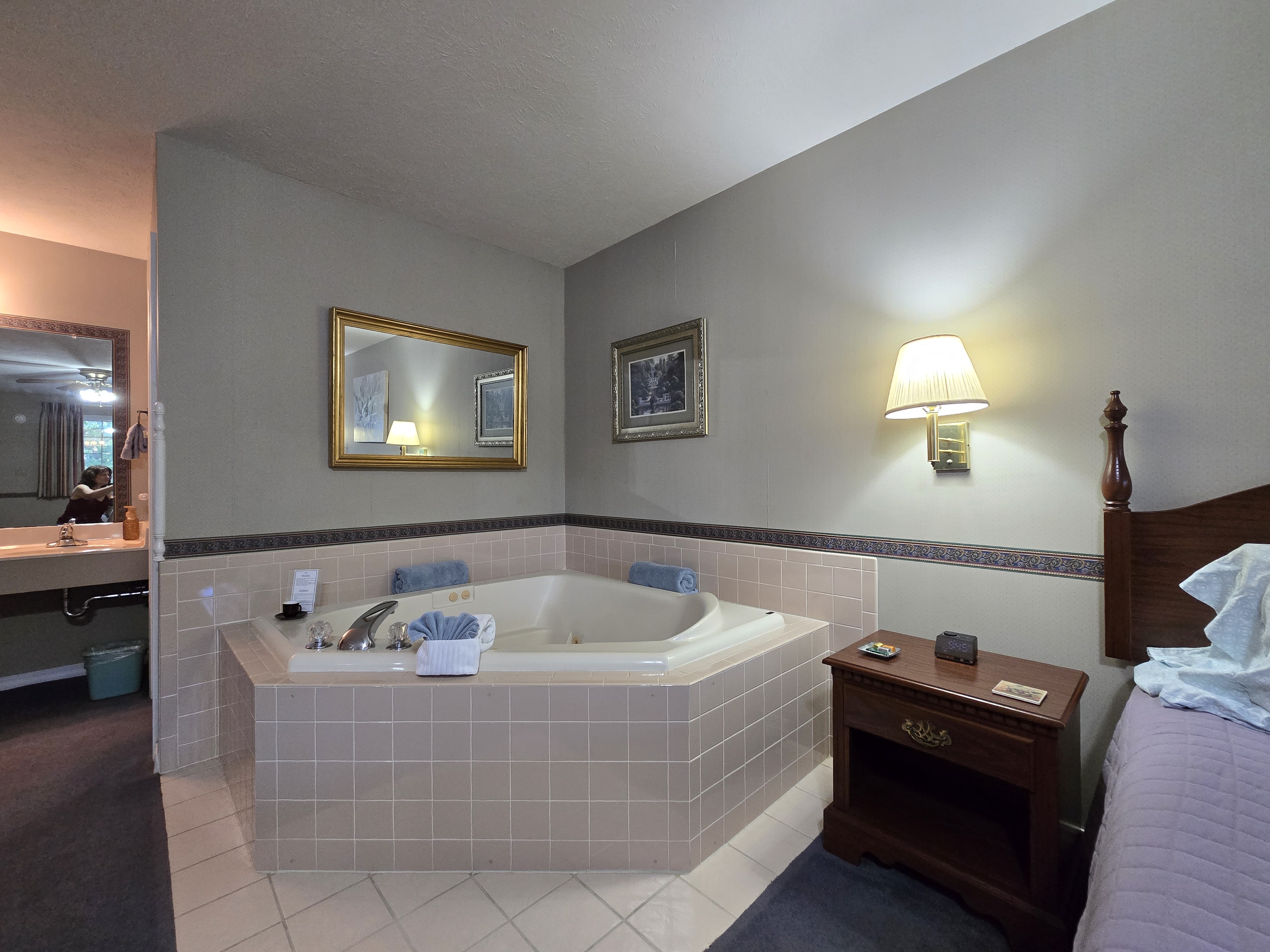Corner view of an octagonal jetted tub with light pink tiled surround, next to a nightstand, with a large mirror and artwork on the wall. The sink/vanity area is visible to the left.