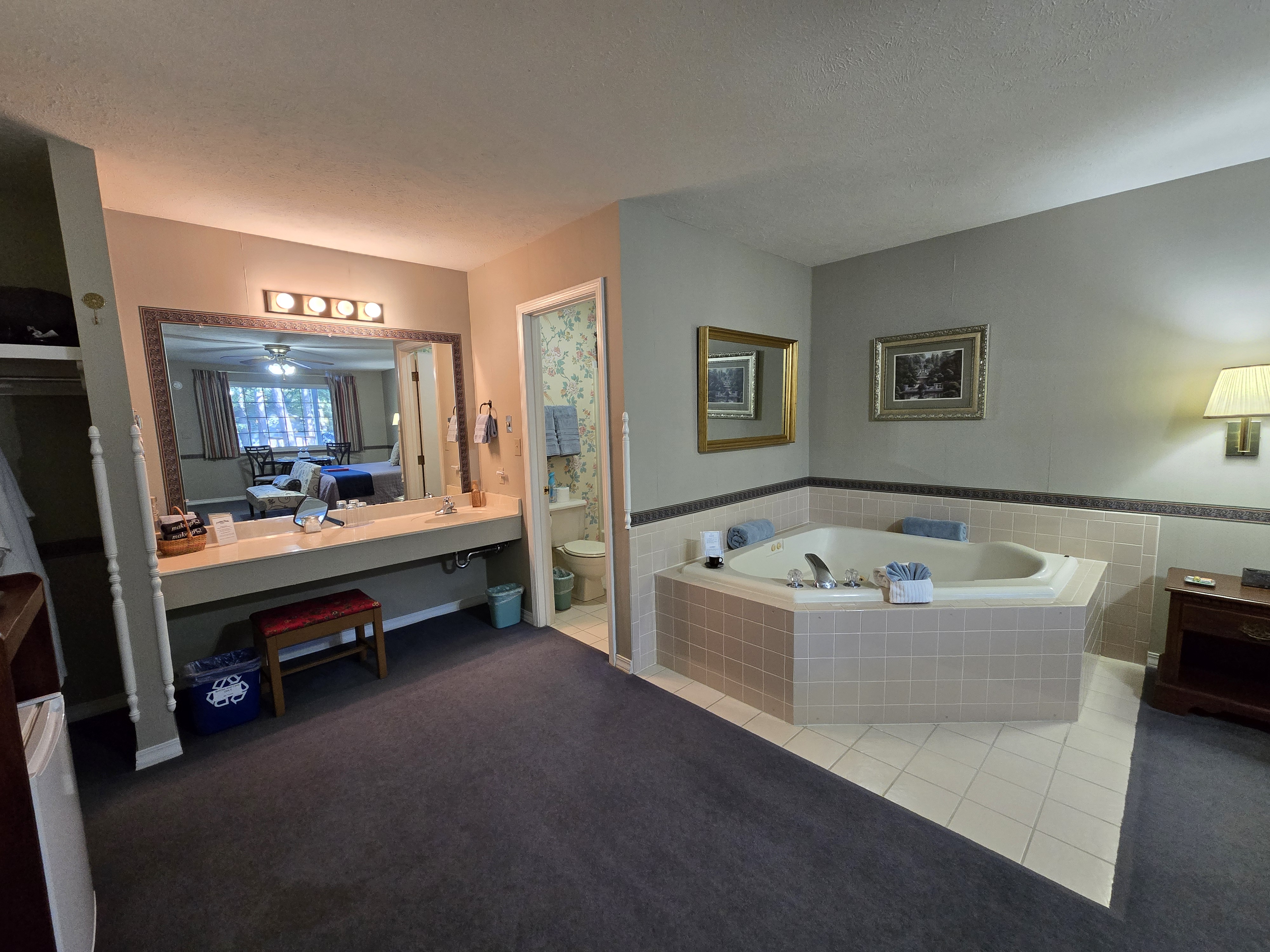 featuring a corner jetted tub with light pink tile surround, a long double vanity with a large mirror, and a doorway leading to a small toilet/shower room.