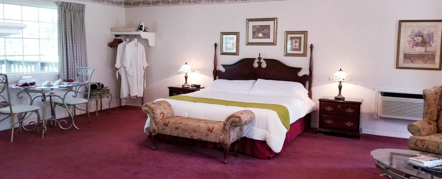 View of the foot of the King sized bed with a wooden headboard. White bedding is shown with a sage green bed scarf and a bench at the end of the bed. The carpet is a maroon color. 3 pictures are on the wall above the headboard and there are nightstands with lamps as well. There is a large back window looking out to the secluded woods on the left of the picture. In front of the window sits a glass, 2-person table with 2 iron chairs. There is an open-style closet space by the table with 2 white, waffle robes hung on 2 of the hangers.