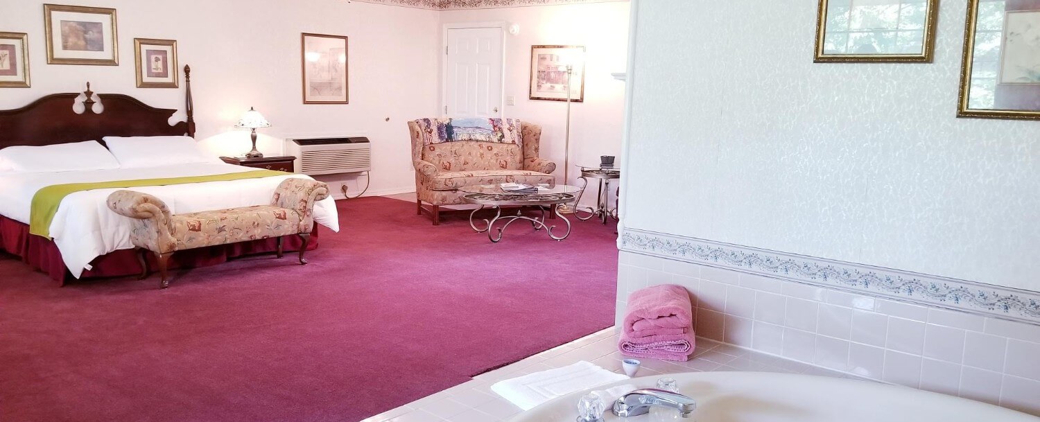 View across the room where you can see the front exit door, a floral loveseat with a coffee table in front, and the king bed with bench at the end. The carpet is maroon and the walls are tan.