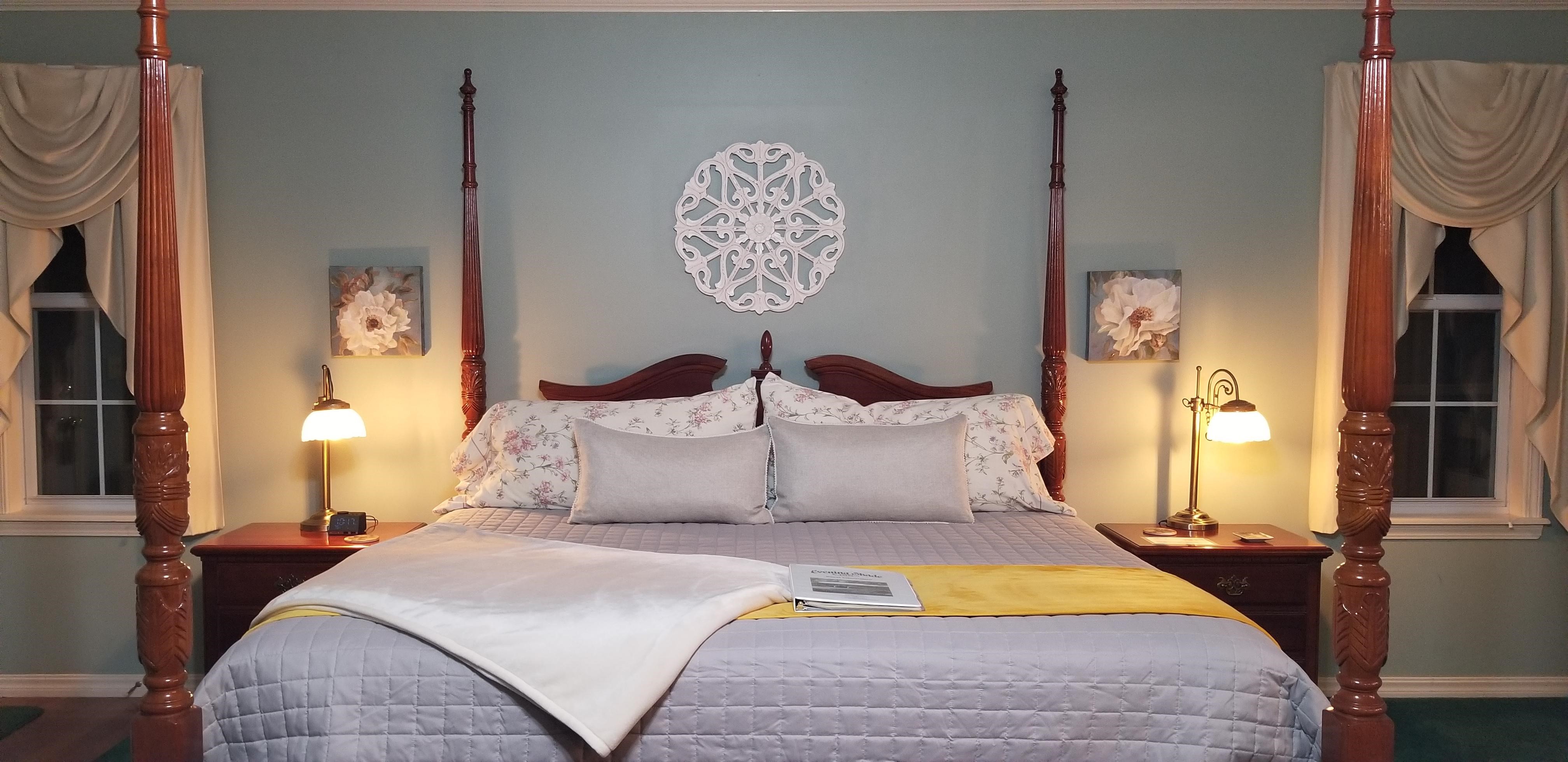 Interior room view with king sized bed as the main focal point. Bedspread is a light grey and has a folded soft, velvet cream blanket at the foot of the bed on the left. There is a yellow, velvet bed scarf at the foot of the bed. Bed is a wooden, classic style, 4 poster bed. The walls are a pale blue. There is a wooden mandala on the back wall above the headboard in the center of the bed. There are bed tables with a lamp on either side of the bed with a window on either side as well.