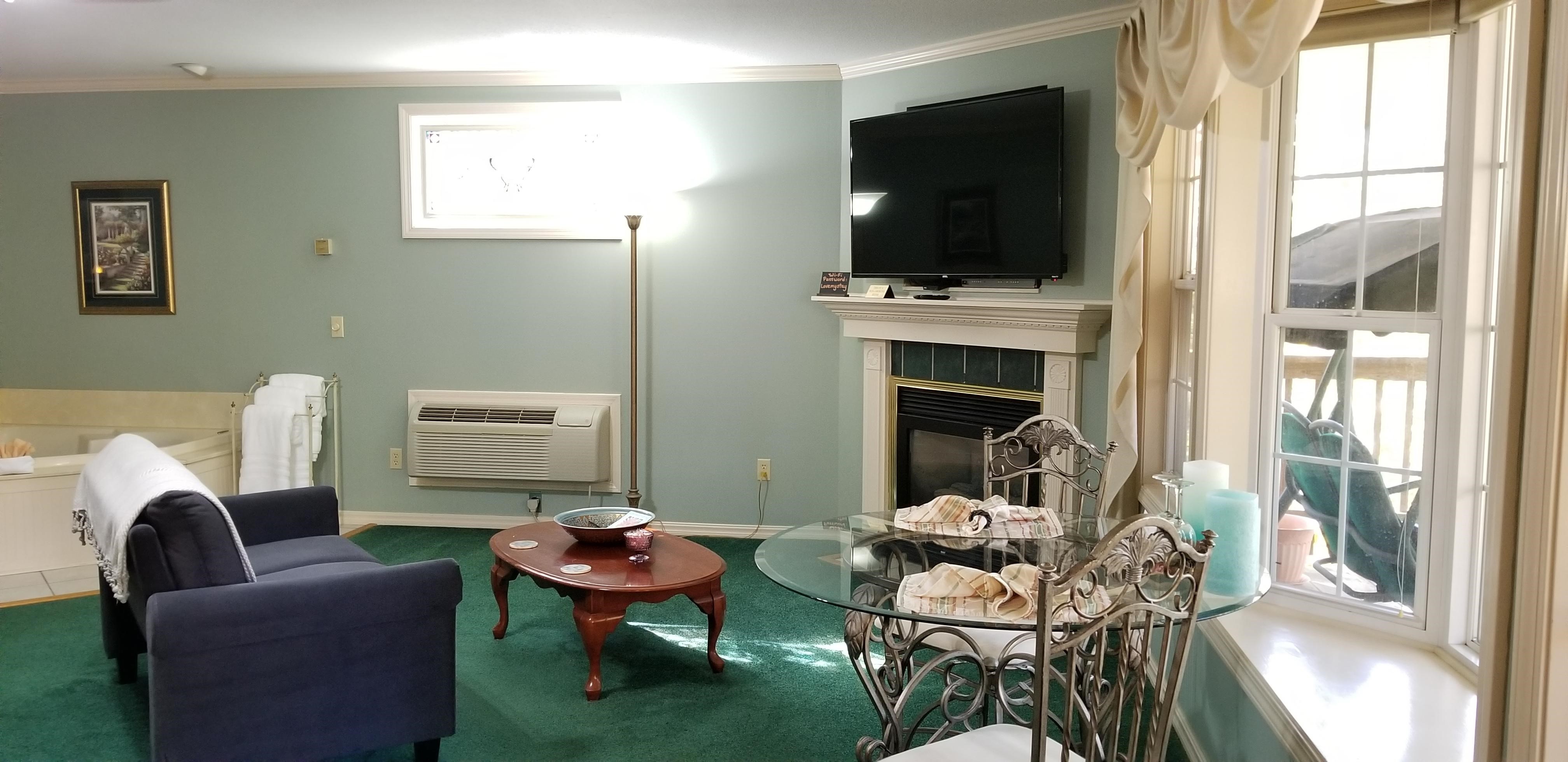 Corner space of interior of room with light blue walls and dark green carpeting. There is a fireplace that is gas in the far right hand corner that has a white surround and mantle piece. There is a flat screen TV above the fireplace on the wall. In the middle of the image is a wooden coffee table with a blue velvet loveseat next to it. The loveseat and coffee table face the fireplace and TV. To the right of the image is the 2 person dining table with glass top and bay window.