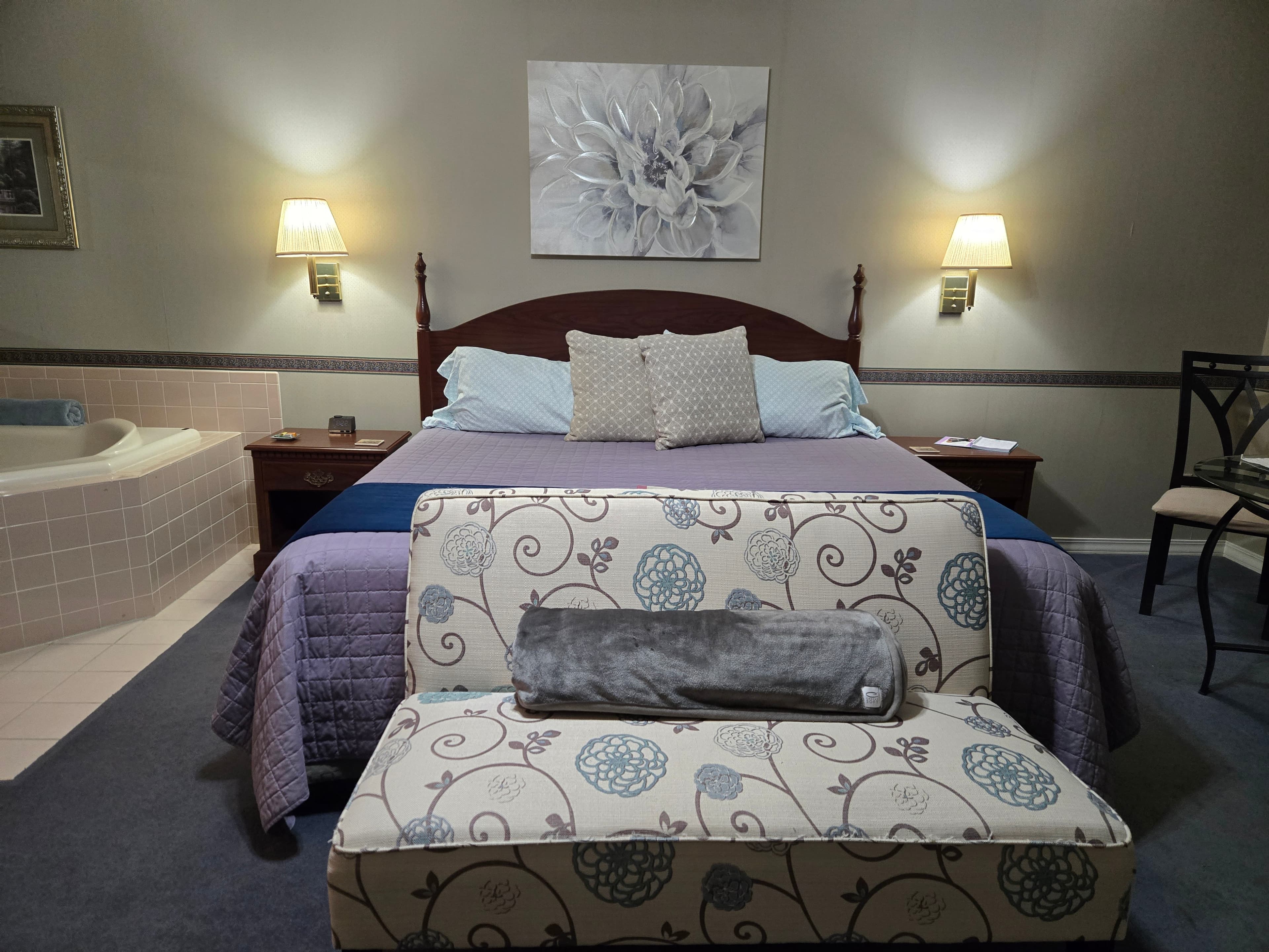 In-room photo taken from the foot of the bed. The walls are a gray color with a border of wallpaper at mid room level. The carpeted flooring is a Taupe color. Ther eis a king size bed with a purple bedspread, blue overlay at the end, and a floral foot bench at the base of the bed. on the wall and a wooden nightstand on either side.