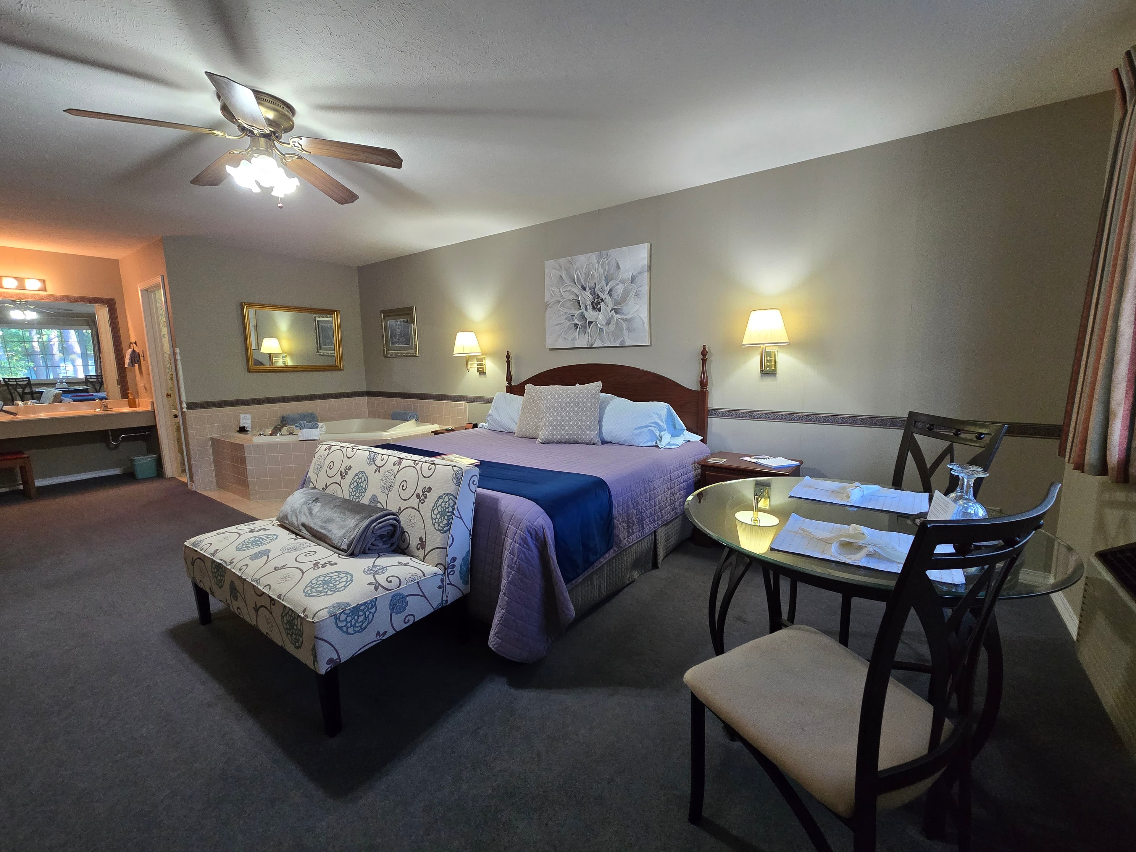 room with a king bed, chaise lounge, small glass dining table, jetted tub in the corner, and a view into the bathroom vanity area.