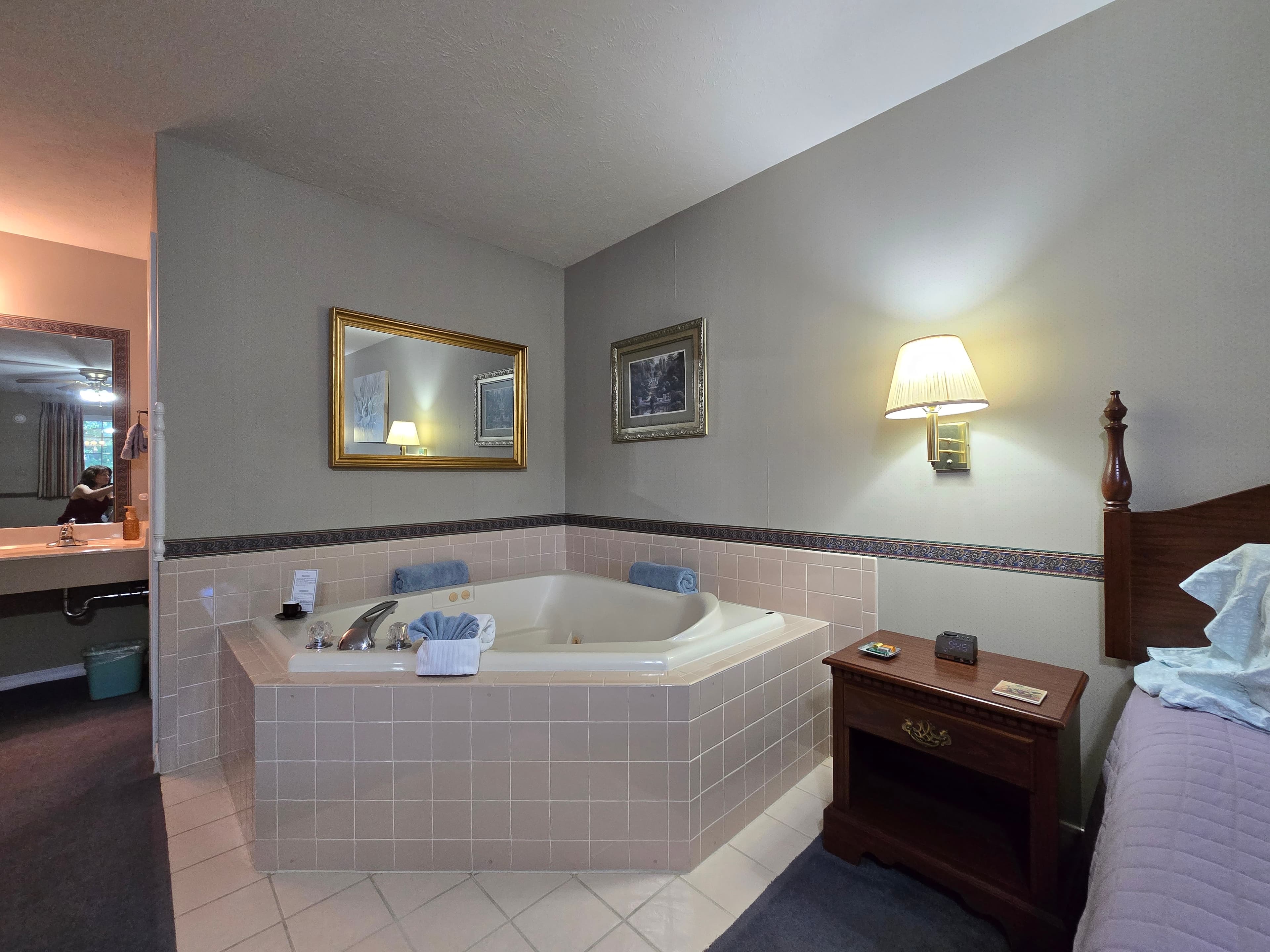 Corner view of an octagonal jetted tub with light pink tiled surround, next to a nightstand, with a large mirror and artwork on the wall. The sink/vanity area is visible to the left.
