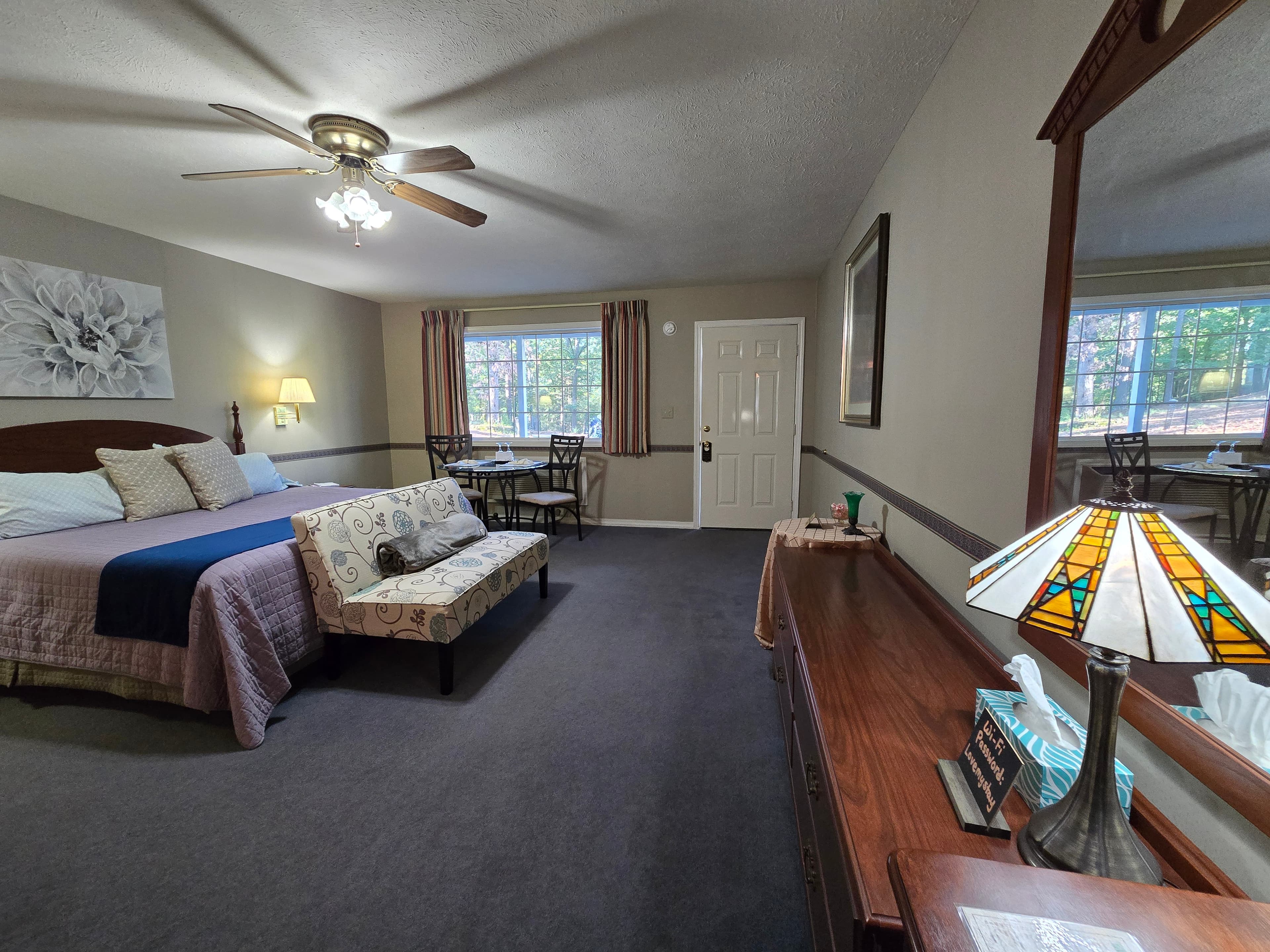 room showing a king bed with a floral art piece above it, a patterned chaise lounge, a small dining table by the window, and a long wooden dresser with a stained glass lamp.