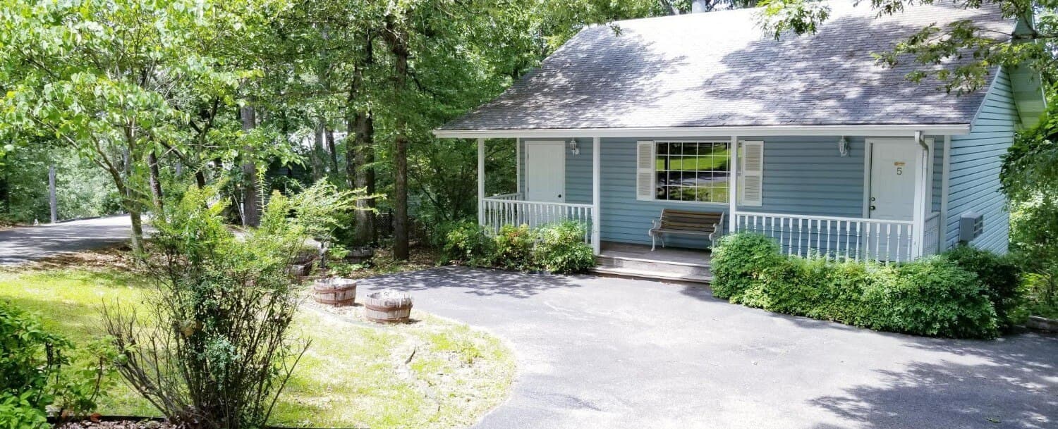 Exterior, detached, light blue, cottage photo. There is a front porch with white wooden rails and a wooden bench. Behind the bench is a large front window. In front of the cottage is the private, paved parking. There is a small yard space in the front left with grass, some trees on the sides of the picture, and bushes in front of the front porch.