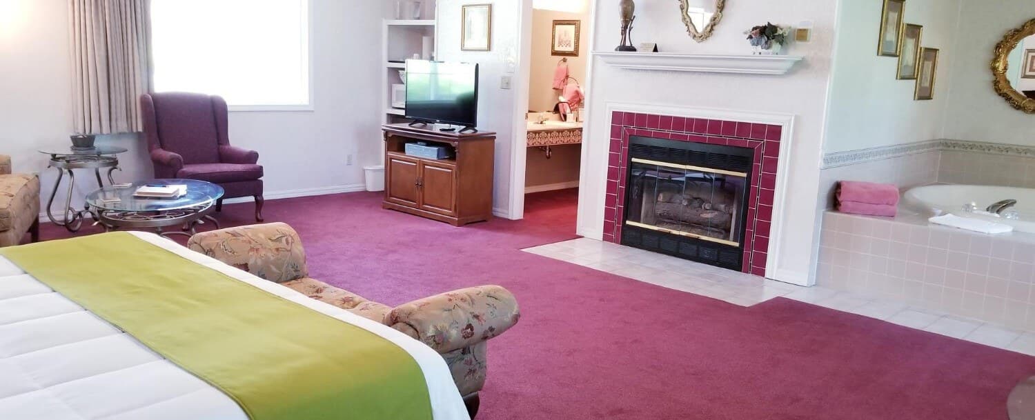 View of the foot of the King sized bed. White bedding is shown with a sage green bed scarf and a bench at the end of the bed. The carpet is a maroon color. Across from the bed is a gas fireplace on the wall. To the left of the fireplace is entry to the bathroom and to the left of that is the flat screen TV. To the right of the fireplace is the 2-person, jetted tub. In the far, back, left of the photo is a large window with a wing backed chair in front and a glass coffee table.