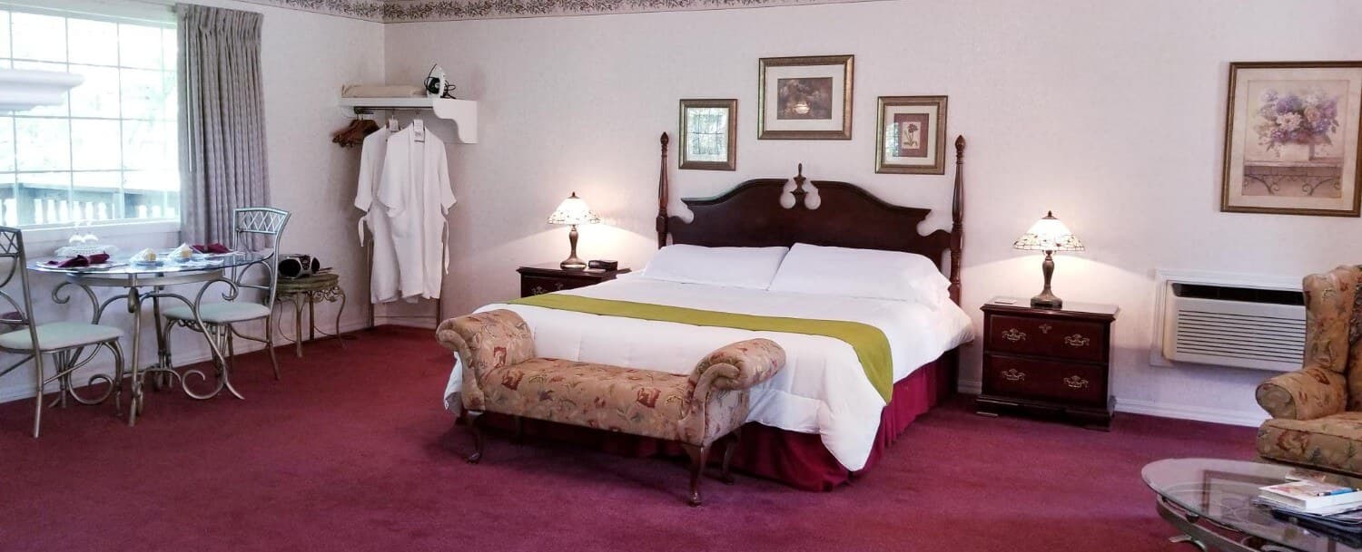 View of the foot of the King sized bed with a wooden headboard. White bedding is shown with a sage green bed scarf and a bench at the end of the bed. The carpet is a maroon color. 3 pictures are on the wall above the headboard and there are nightstands with lamps as well. There is a large back window looking out to the secluded woods on the left of the picture. In front of the window sits a glass, 2-person table with 2 iron chairs. There is an open-style closet space by the table with 2 white, waffle robes hung on 2 of the hangers.