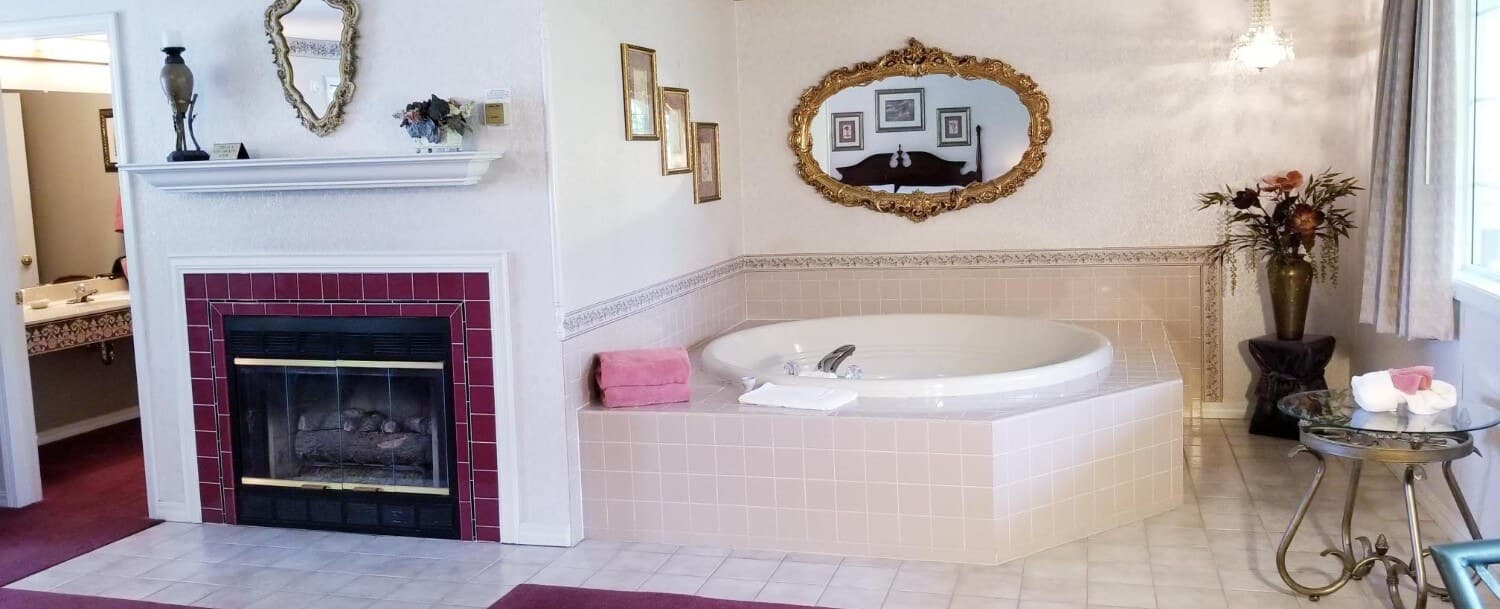 White trimmed, with maroon tile, gas fireplace is shown on the left with a mirror above the mantle. To the right is the 2-person, jetted tub that is large and round. There is a small, round, glass and iron side table to the far right. The walls of the room are pale tan.