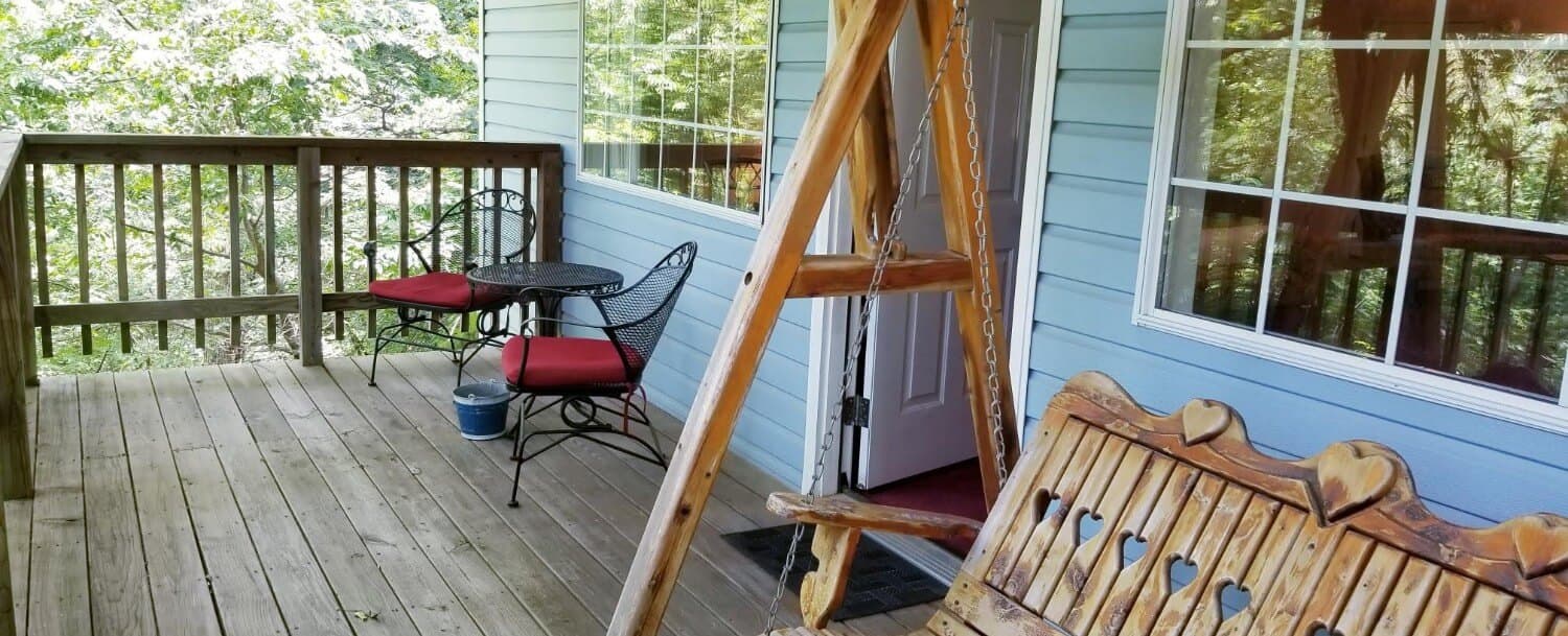 Private wooden back deck that looks into the wood line. 2 person wrought iron table and chairs with maroon cushions. There is a wooden, 2-person porch swing. Some of the exterior, light blue siding shows with it's 2 big windows. You can also see the entry door that goes into the cottage.