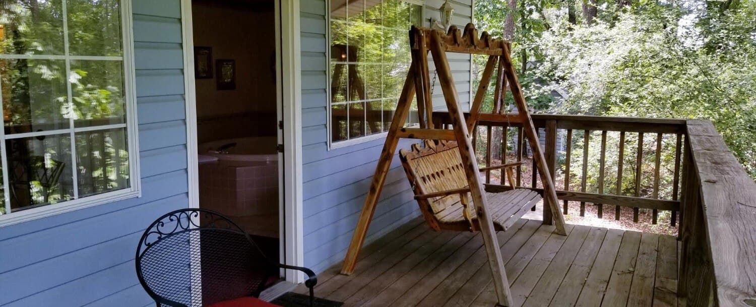 Private wooden back deck that looks into the wood line. There is a wrought iron chair. There is a wooden, 2-person porch swing. Some of the exterior, light blue siding shows with it's 2 big windows. You can also see the entry door that goes into the cottage.