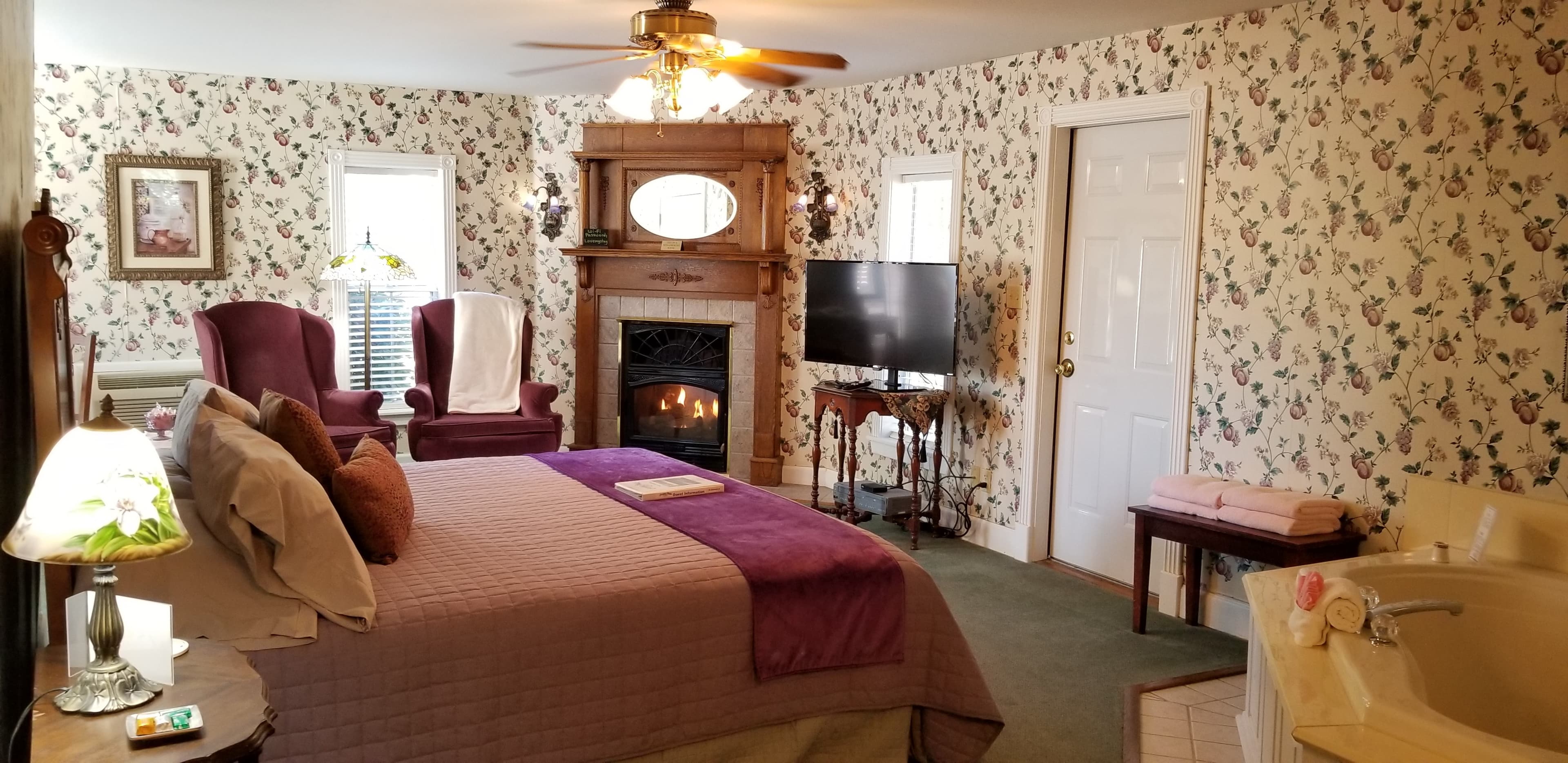Across the interior of the floral wallpapered room. On the far back wall, in a corner, is a lit, gas fireplace in an old fashioned style with an oval mirror sideways embedded in the wood mantle space. To the left of the fireplace on the back wall are 2 armchairs that are purple with a Tiffany floor lamp in between. To the right of the fireplace is a table with the flat screen TV on top. To the right of the TV is a door that goes out to the private back deck. In the foreground is a side view of the king sized bed with a purple bedspread and purple sash at the foot of the bed.