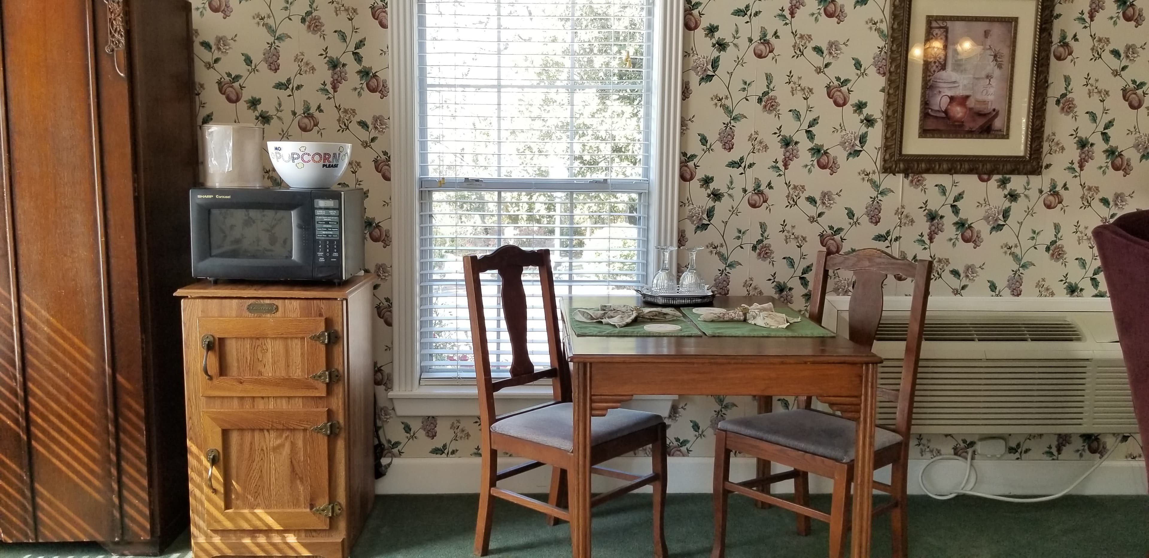 Interior view of the guestroom's dining space. 2 person wooden table dressed with napkins and placemats. There is a window on the wall behind the table. To the left of the table is a mini fridge that looks old fashioned with a wooden-look exterior. On top is a microwave and popcorn bowl. The wallpaper in the room is floral. There is a framed picture on the wall to the right of the table. The carpet is forest green.