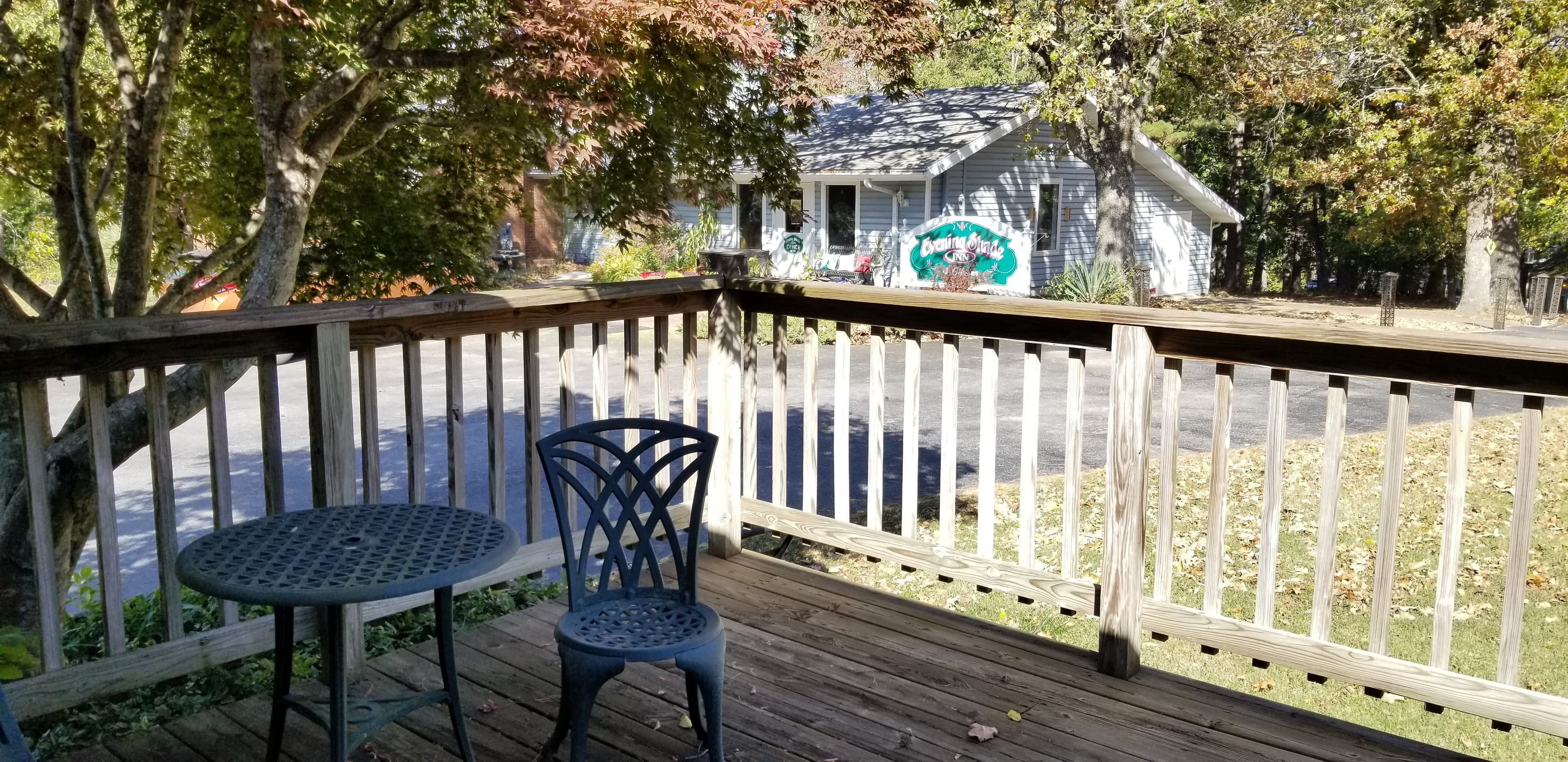 Exterior view of the back deck of Number 6, Magnolia Suite. The view shows the wooden deck with wooden rails. There is a wrought iron cafe table for 2. There is a Japanese Maple tree with it's foliage shading the left side of the deck. In the background is a backyard space and a view of the corner of the office building on the grounds.