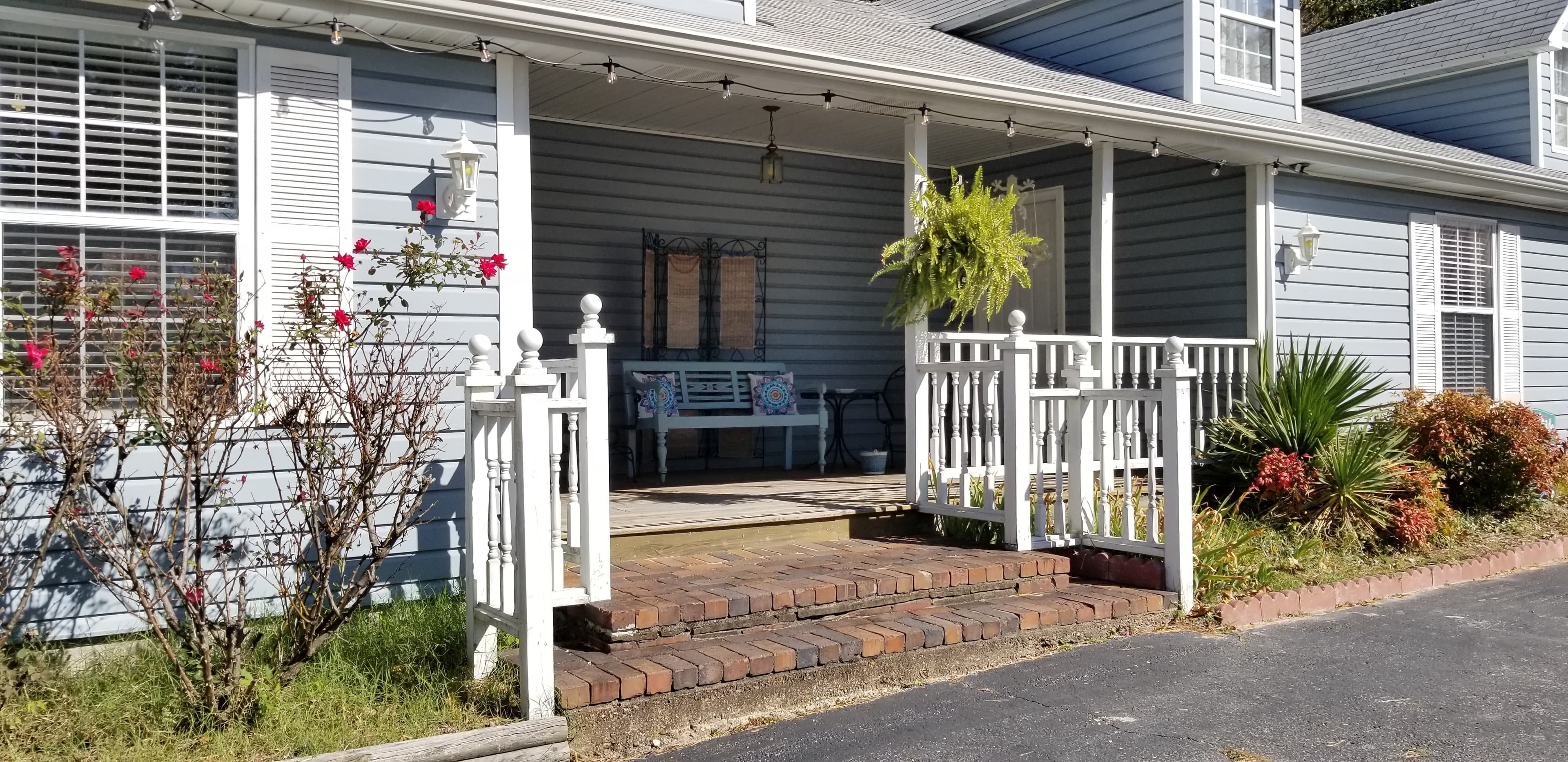 Exterior, front view of the building that houses both the Magnolia and Gardenia Suites. The siding is a light blue and the trim and railings are white. There are a few brick stairs leading up to the deck in the front that sits between the entry to each room. There are dormers shown at the top of the building. On the deck in the middle is a blue bench. There is a fern hanging out front and garden beds with flowers and plants on the sides of the front of the building. The exterior windows on the building have the white trim and are flanked with shutters in white.