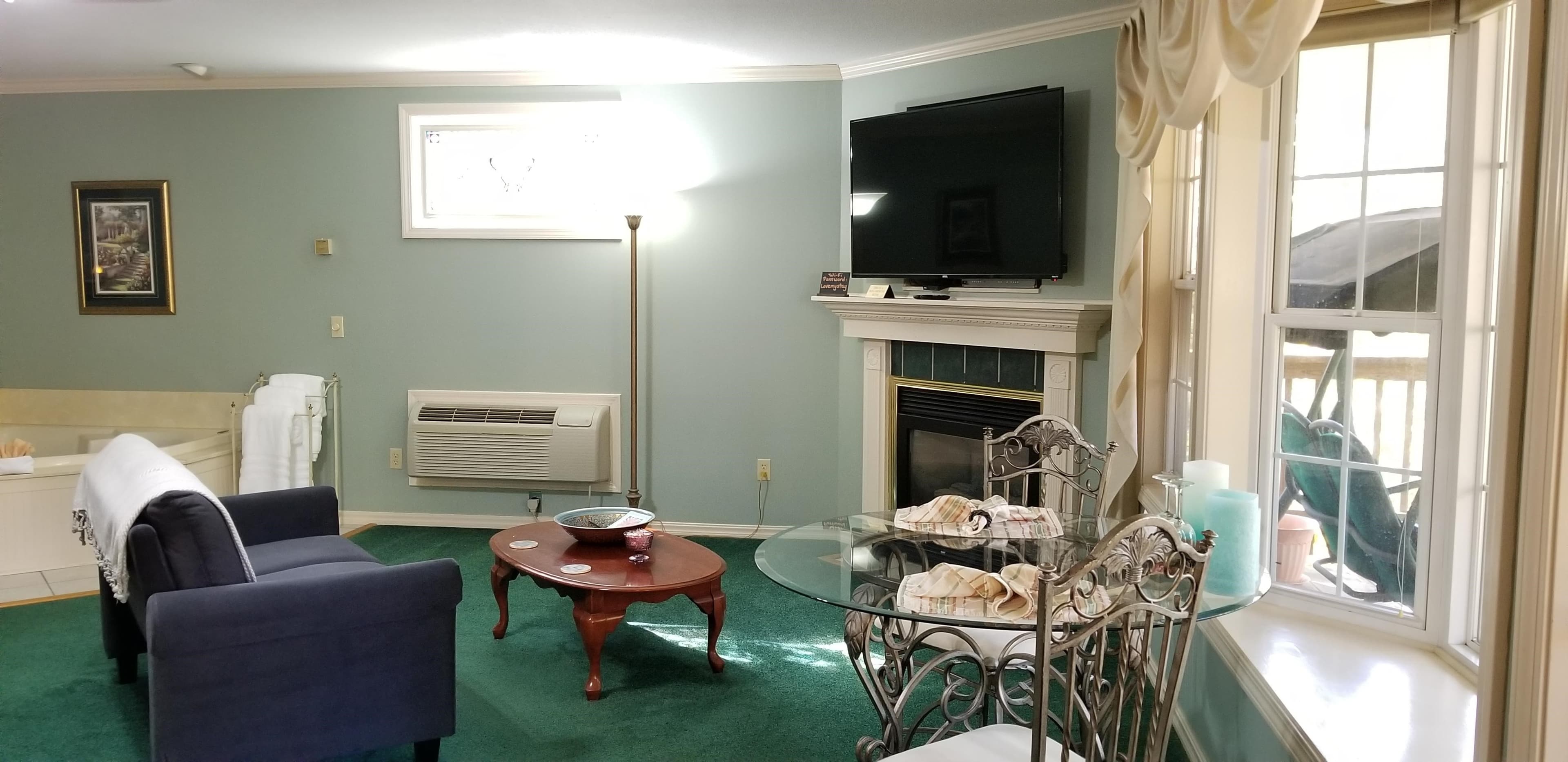 Corner space of interior of room with light blue walls and dark green carpeting. There is a fireplace that is gas in the far right hand corner that has a white surround and mantle piece. There is a flat screen TV above the fireplace on the wall. In the middle of the image is a wooden coffee table with a blue velvet loveseat next to it. The loveseat and coffee table face the fireplace and TV. To the right of the image is the 2 person dining table with glass top and bay window.