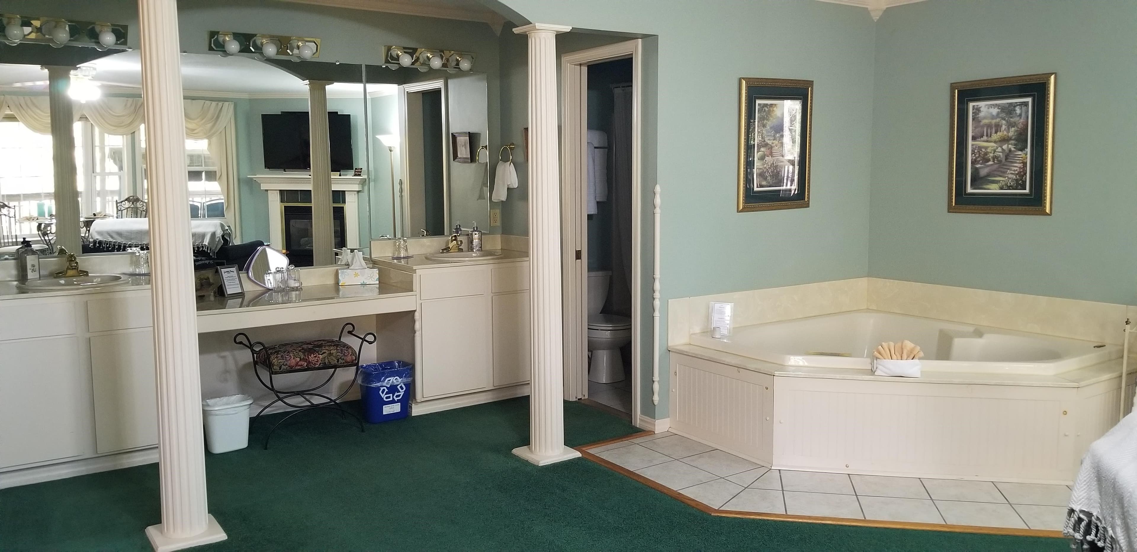 Interior view looking toward the vanity/bathroom/jetted tub. Jetted tub and bathroom and vanity are the focal point. The walls are a light blue. There is a seat at the vanity with a floral fabric. The vanity area is recessed and has pillars in front in the arched doorway. The pillars and room trim are all in white. The Carpet is green and the walls are light blue. The jetted tub is in a corner and is tan in color with matching tan surrounding tile on the floor. There is a picture that is framed above either side of the tub.