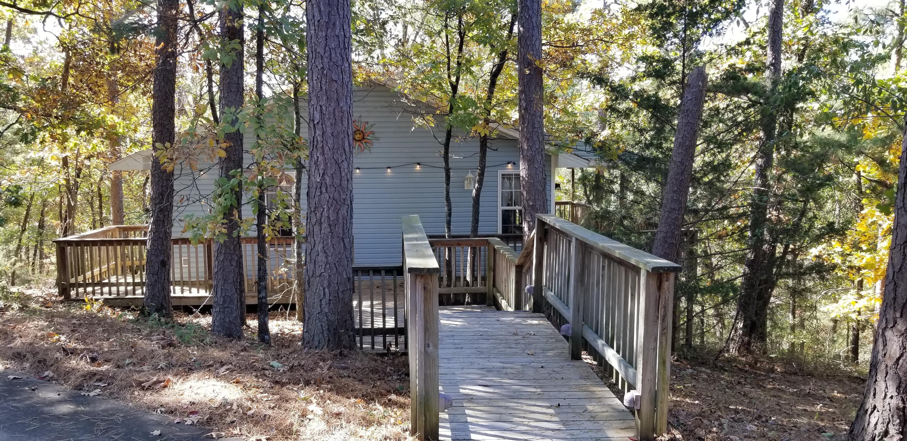 Exterior view of Dahlia Cottage looking across the wooden decking that leads to the cottage. Exterior siding is a light blue. There are woods surrounding the cottage and a string of large bulb cafe lights on the front of the building. Above the string of lights is a large decorative metal sun that hangs beneath the peak of the roof on the wall.