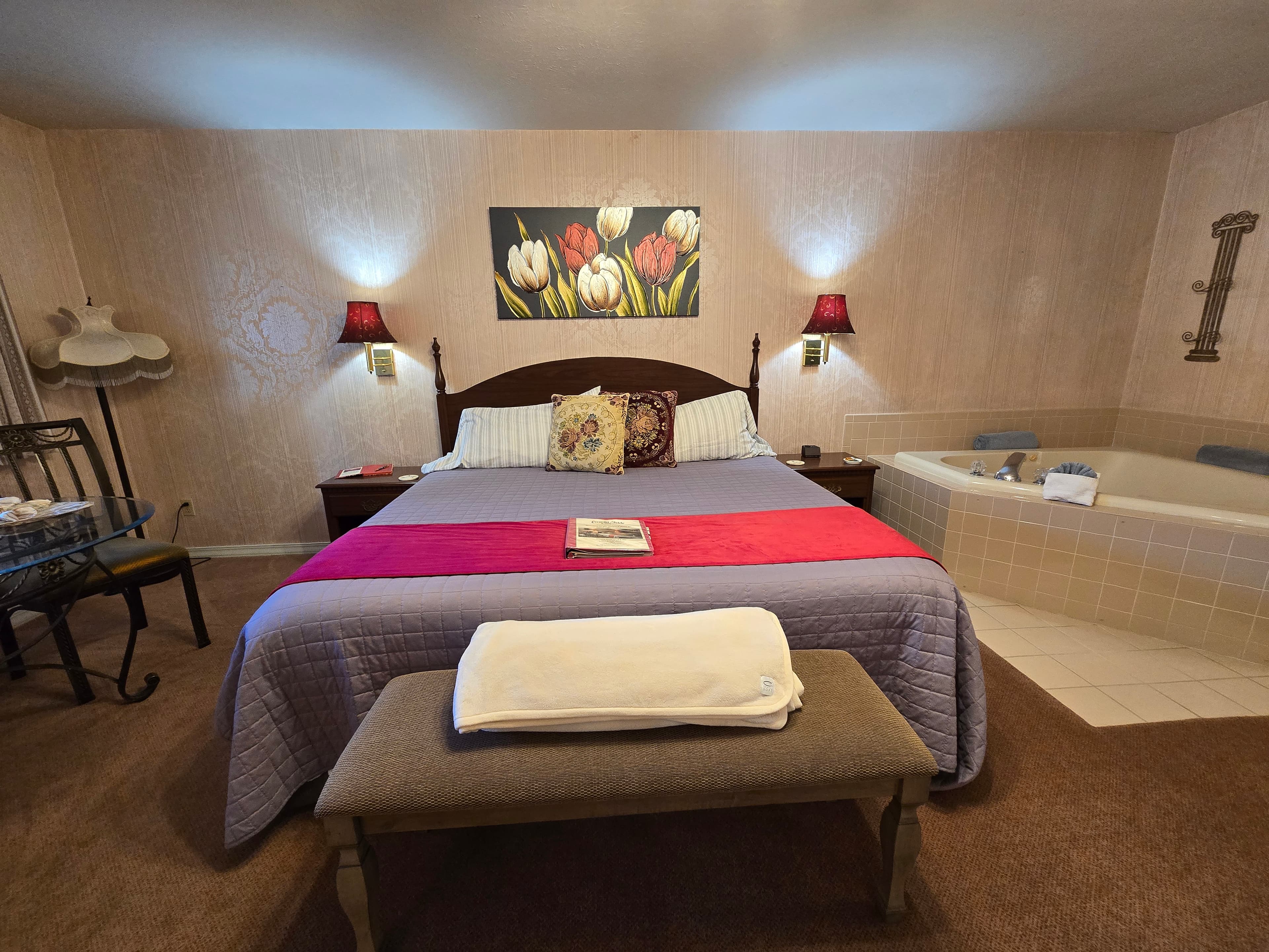 Spacious, well lit, open room with king sized bed. Two-person jetted tub is next to the bed. There are carpeted floors and sconces that cast a warm glowing light.