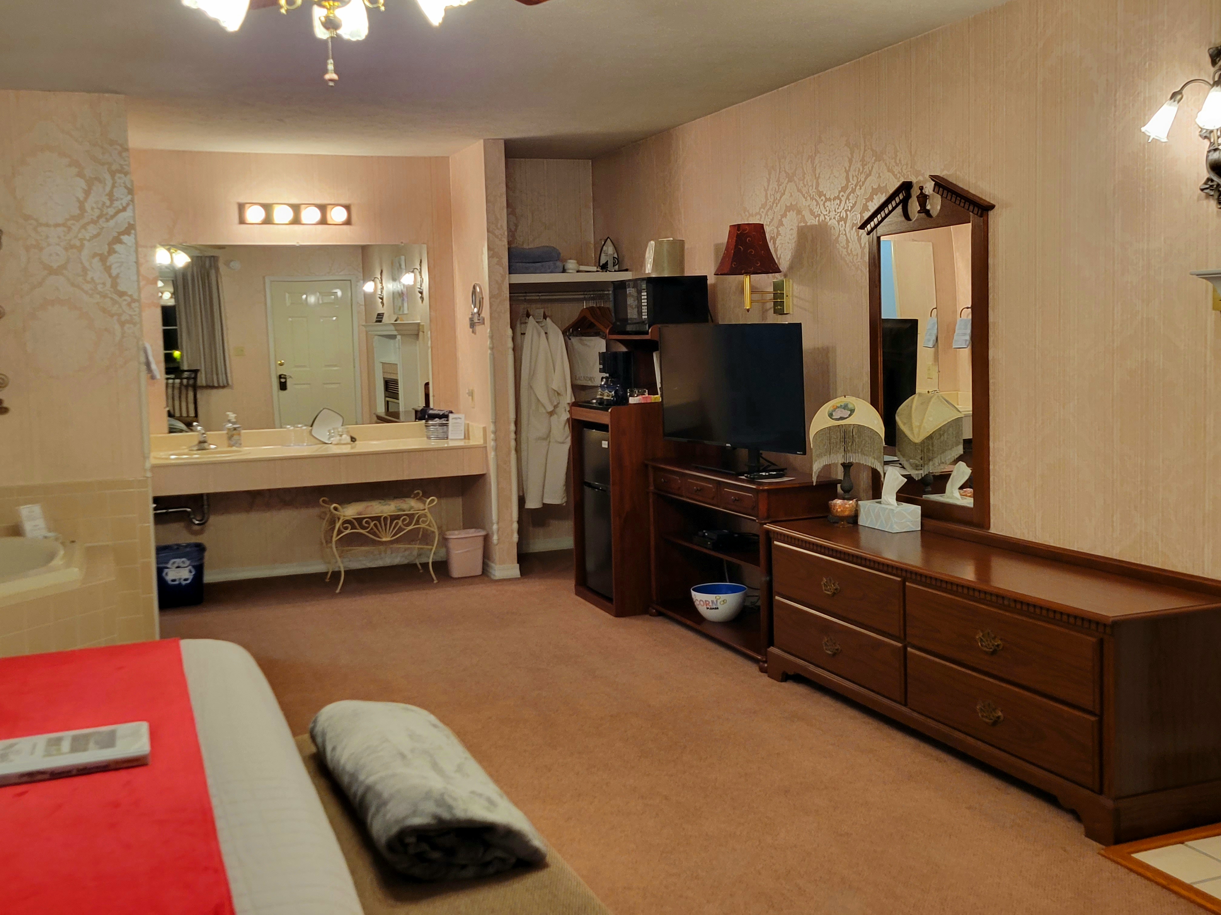 Flat screen TV, elongated four drawer dresser with a mirror above it, coffee stand with microwave on top, mugs, coffee pot, sweeteners, glasses, and a fridge is in the lower part of the stand. There is an opened closet space with robes hanging inside.