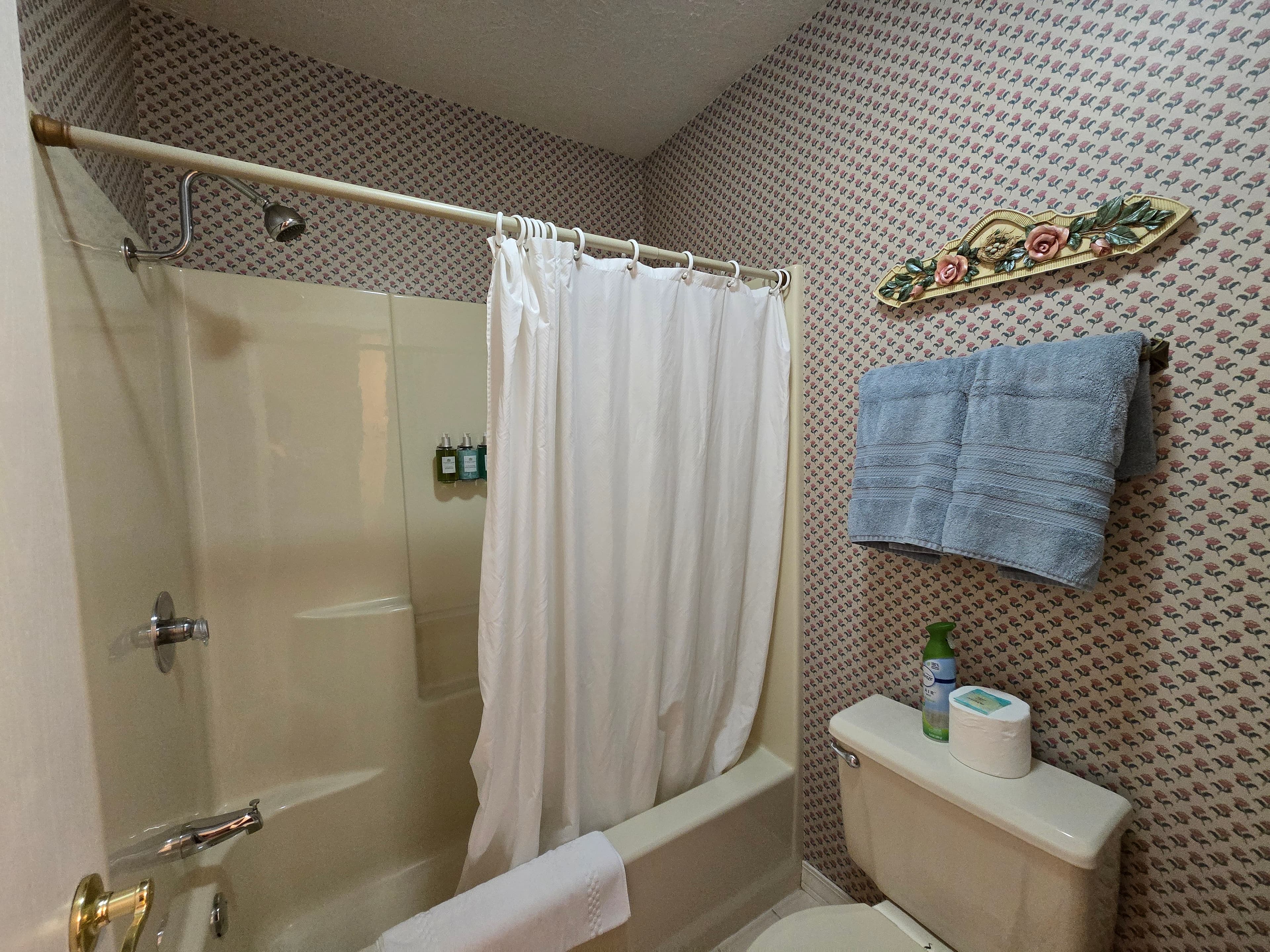 Small bathroom showing a tub/shower combination with a white curtain, toilet, and patterned wallpaper.