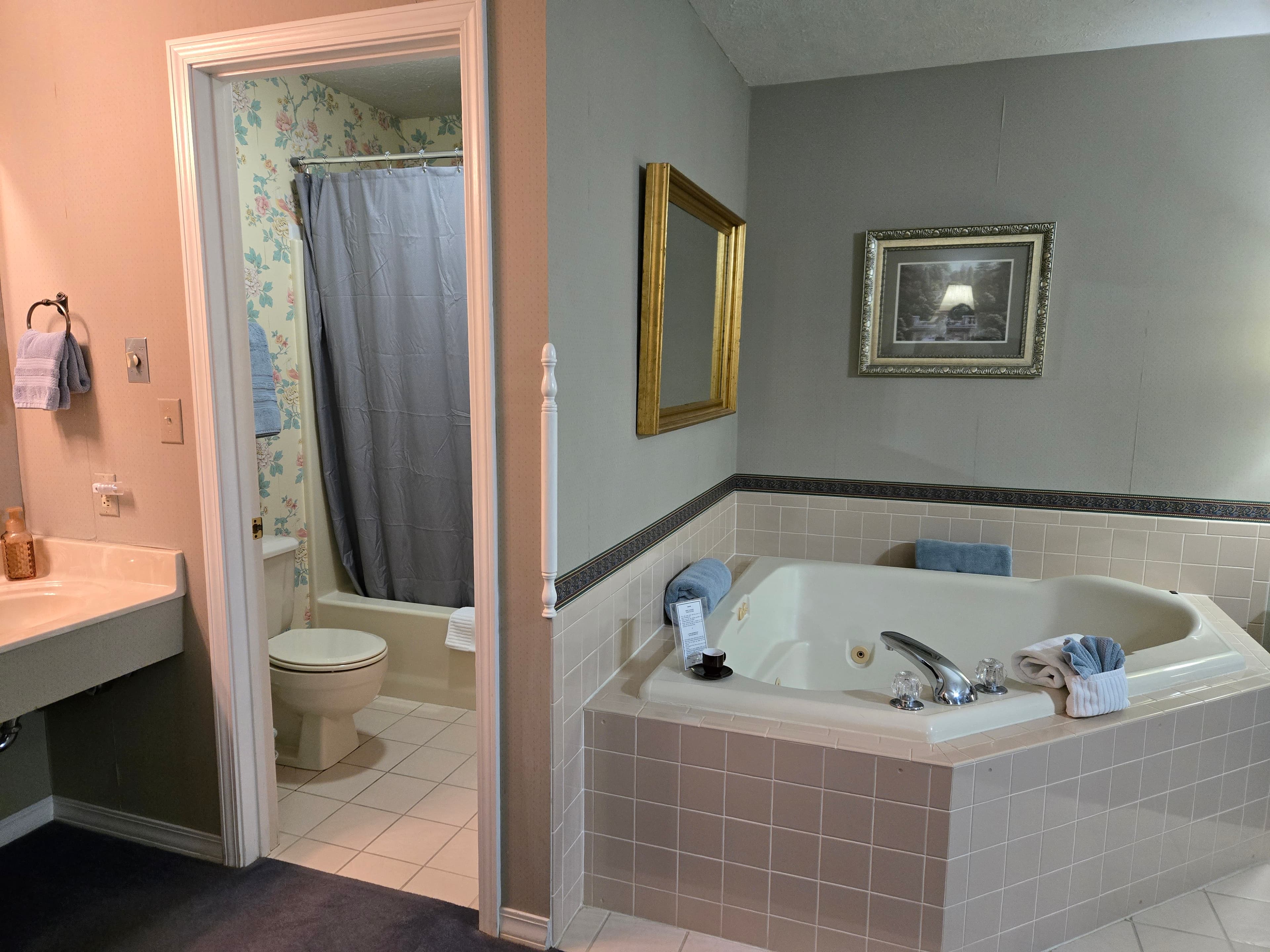 An octagonal jetted tub tiled in light pink, positioned next to an open doorway that shows a small room with a toilet and shower with floral wallpaper. A sink and vanity are visible to the left.