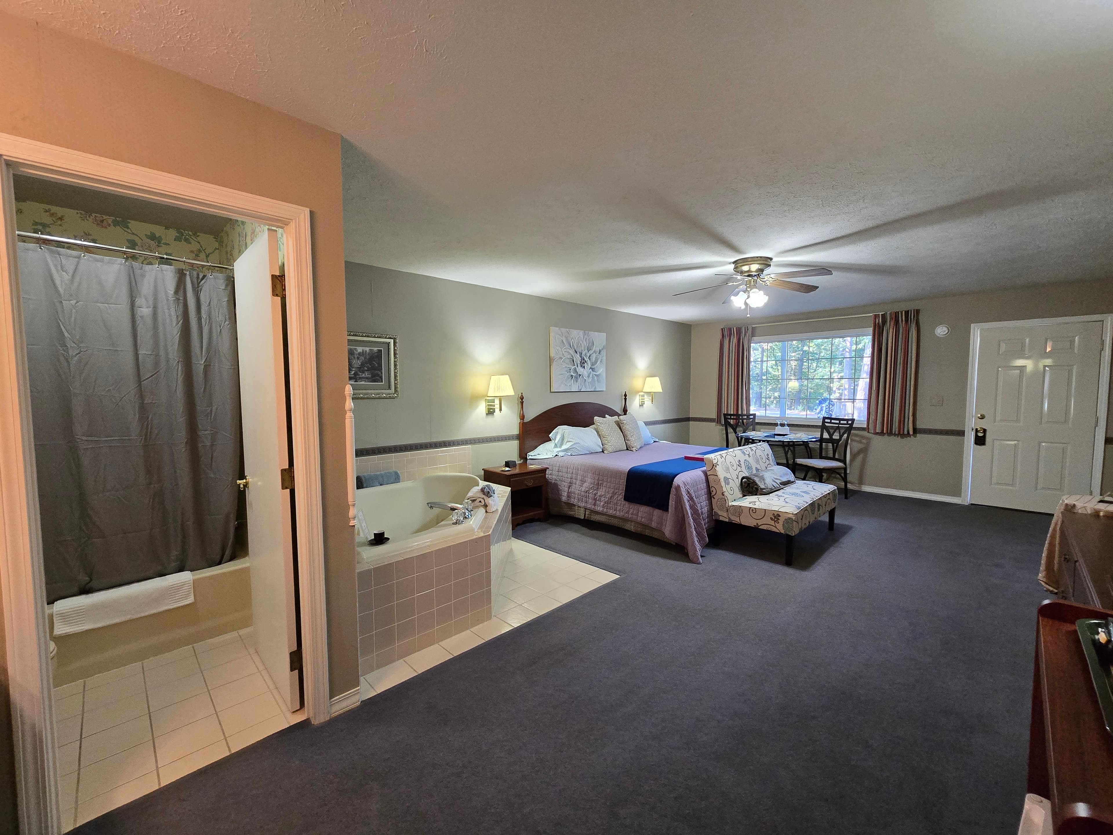 Wide view of a hotel room showing a king bed, a tiled corner jetted tub, a dining table by the window, and an open doorway revealing a small toilet/shower room.