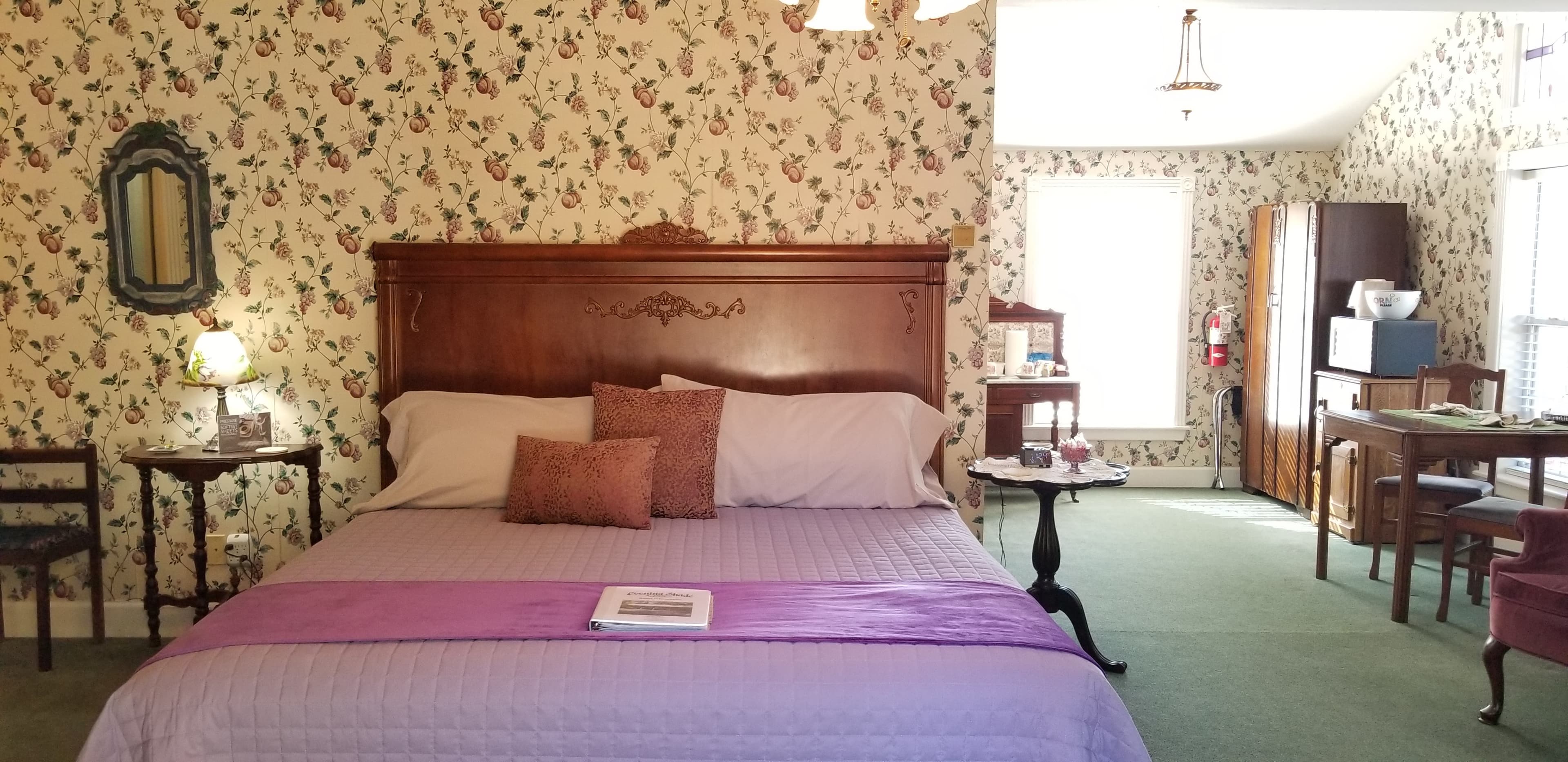 Interior room view with king sized bed, purple bedspread with accent throw pillows. The walls of the room have floral wallpaper. To the right side of the bed is an area of the guestroom that extends to the back of the guestroom where there is a dining table, an armoire, and a window. The carpet in the room is forest green. There are nightstands flanking both sides at the top of the bed.