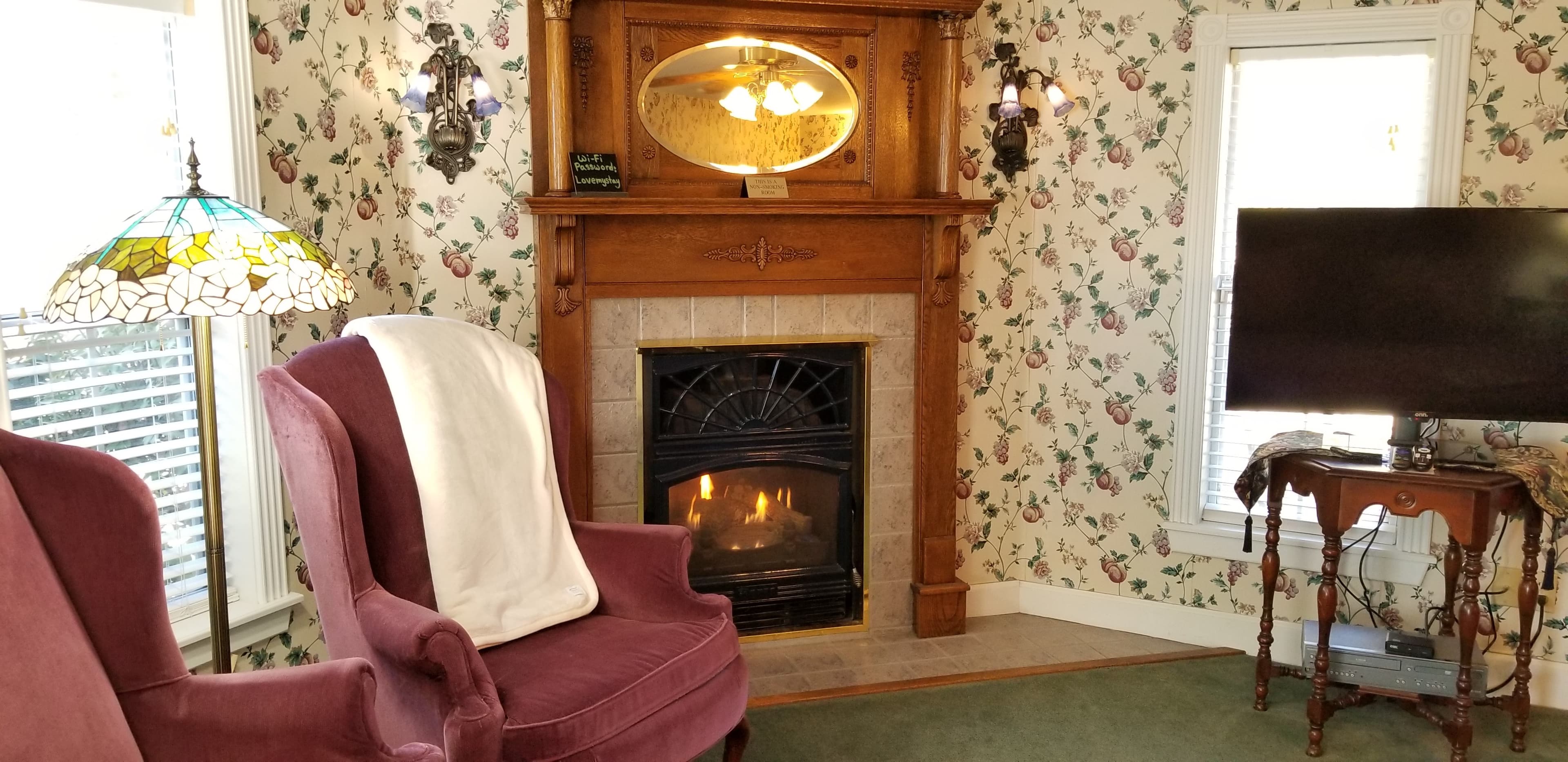 Interior view of room with floral wallpaper and lit gas fireplace in the corner. The fireplace is flanked with fluted, blue sconces. There are 2 purple chairs in the foreground with a Tiffany floor lamp in between. The carpet is a forest green color and there is tile on the floor that is tan in front of the fireplace. To the far right is a flat screen TV on a decorative wooden table or stand.