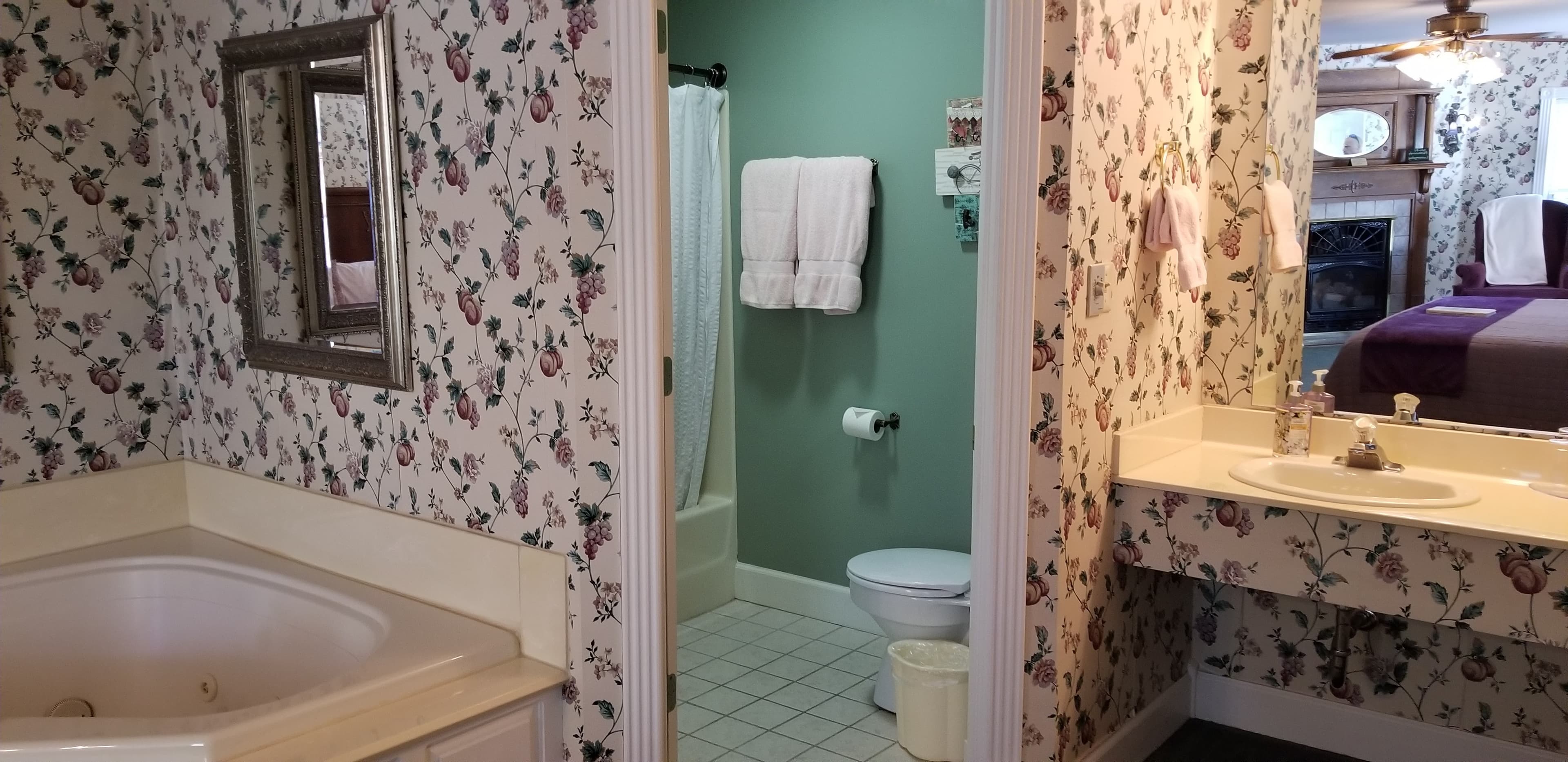View looking into the bathroom space that features a toilet and a shower curtain where the shower/tub combo is in the room. The water closet is painted teal and clean. The corner of the vanity right outside the water closet is also visible and to the left of the water closet is a view of the 2 person jetted tub with framed mirrors on the wall above it. There is floral wallpaper surrounding the vanity and on the walls by the jetted tub.