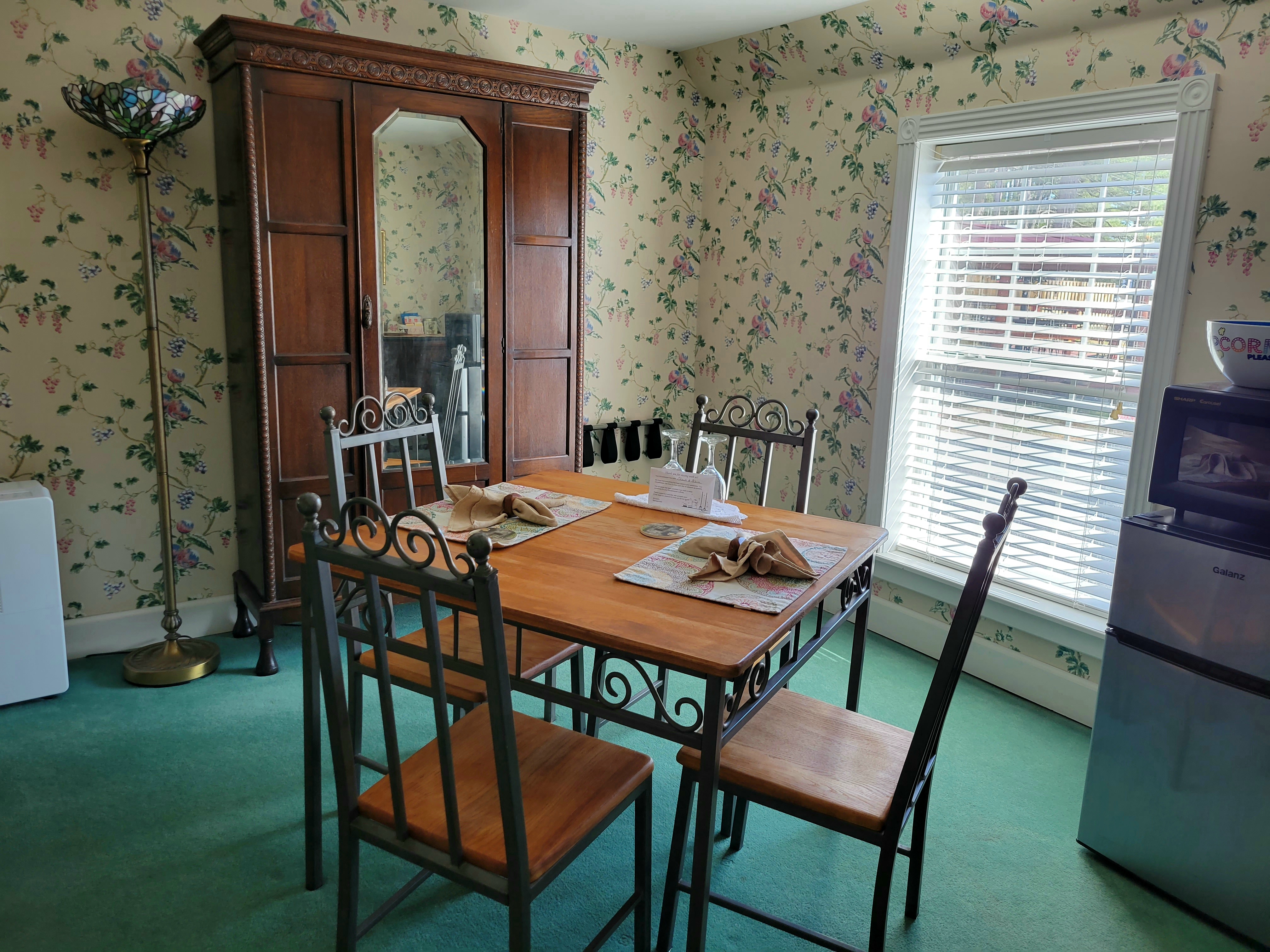 A dining area in a suite featuring a wooden table with four metal chairs set against floral wallpaper. The room includes a large wooden armoire with a mirrored door and a window with white blinds.