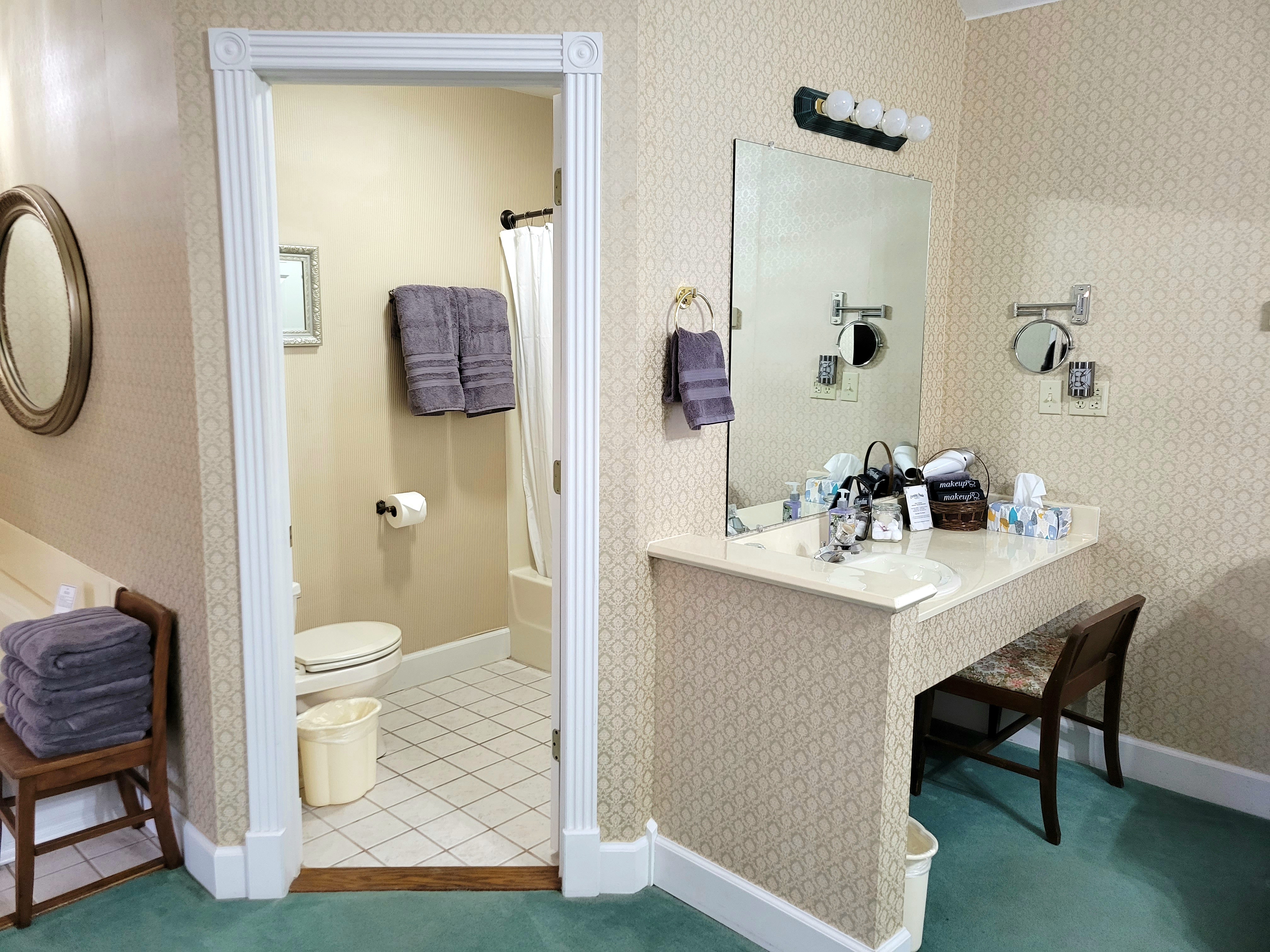 A bathroom featuring a separate vanity area with a large mirror and seating. A doorway leads into a white-tiled bathroom containing a toilet and a shower-tub combination with a white curtain.