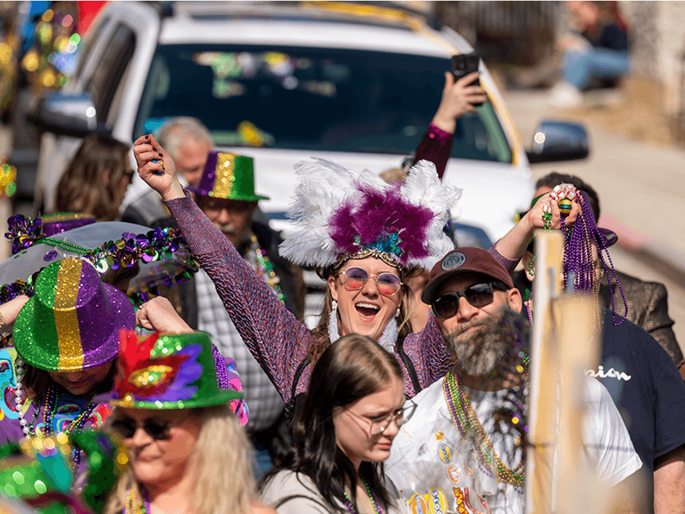 A joyful crowd celebrates a colorful parade. A woman wears a feathered hat with arms raised, surrounded by people in vibrant costumes and beads. Festive and lively atmosphere.