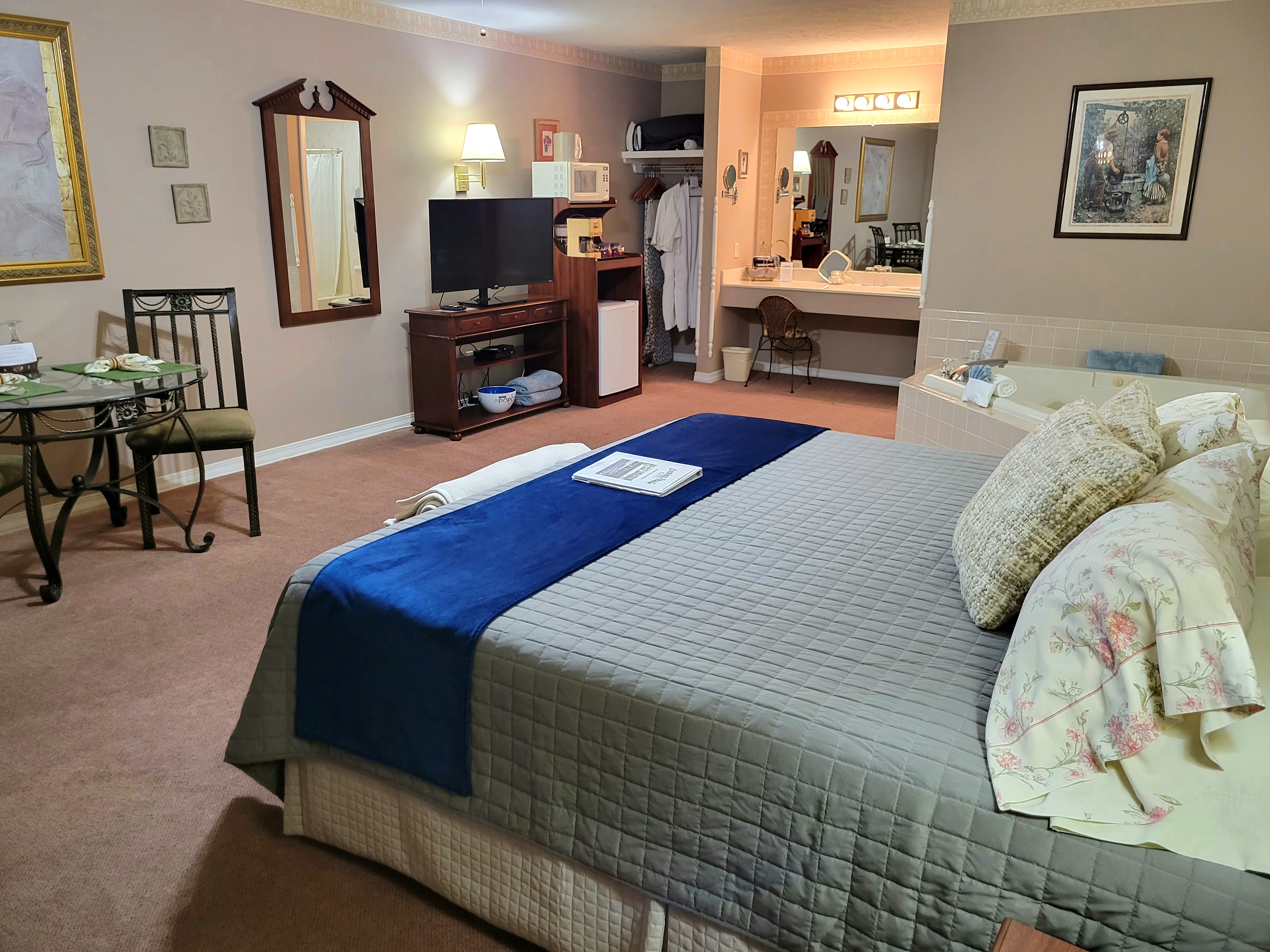 A spacious suite showing a king-size bed with a blue runner, a flat-screen TV on a wooden stand, and a microwave and mini-fridge station. The open-plan room also features a dining table, a large jetted tub, and a vanity area with a desk.