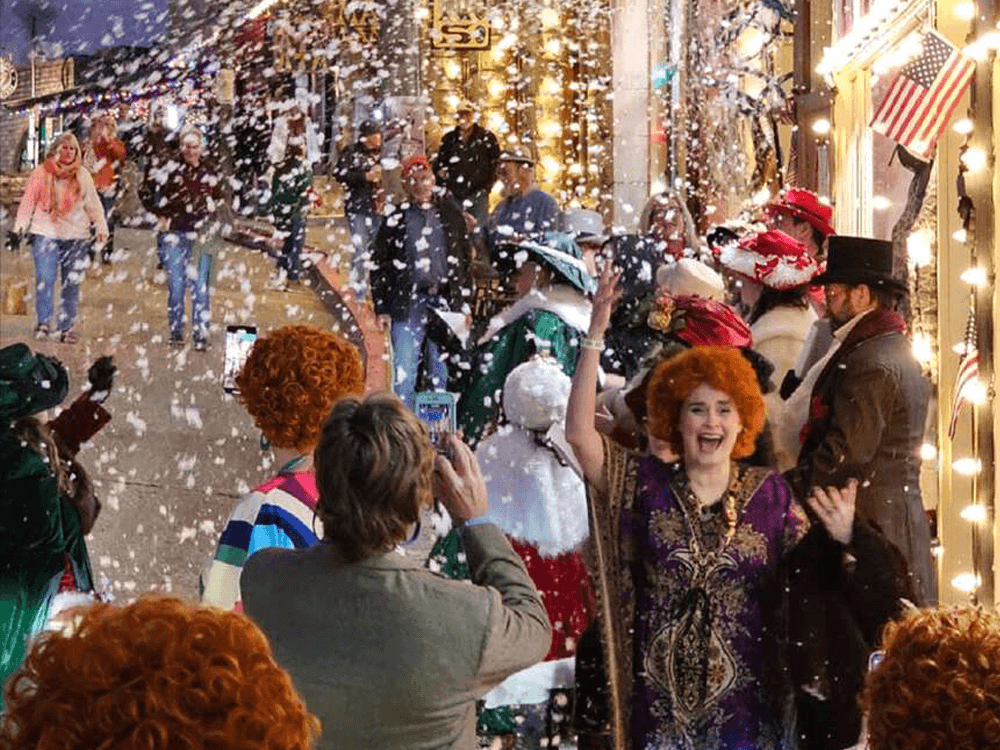 A lively scene of people celebrating in a festive, snow-filled street, with cheerful participants wearing colorful costumes and wigs.