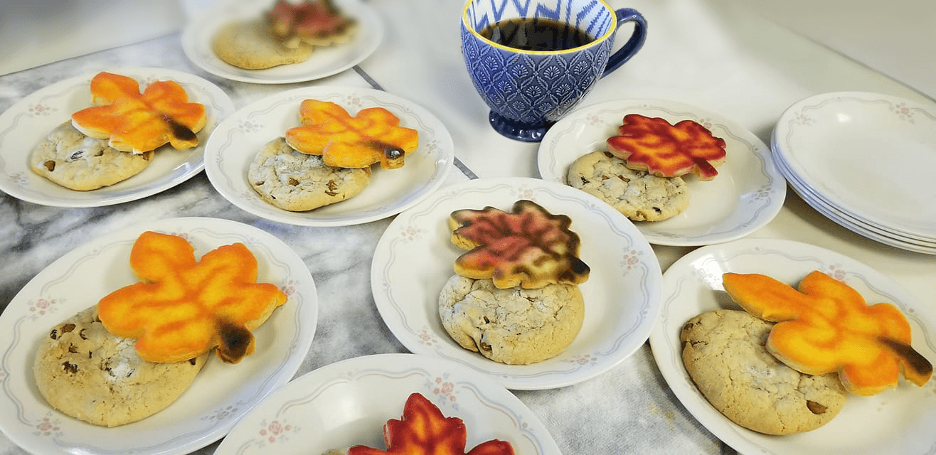 Plates with cookies topped with colorful autumn leaf-shaped cookies. A patterned mug holds a beverage, creating a warm, cozy fall atmosphere.