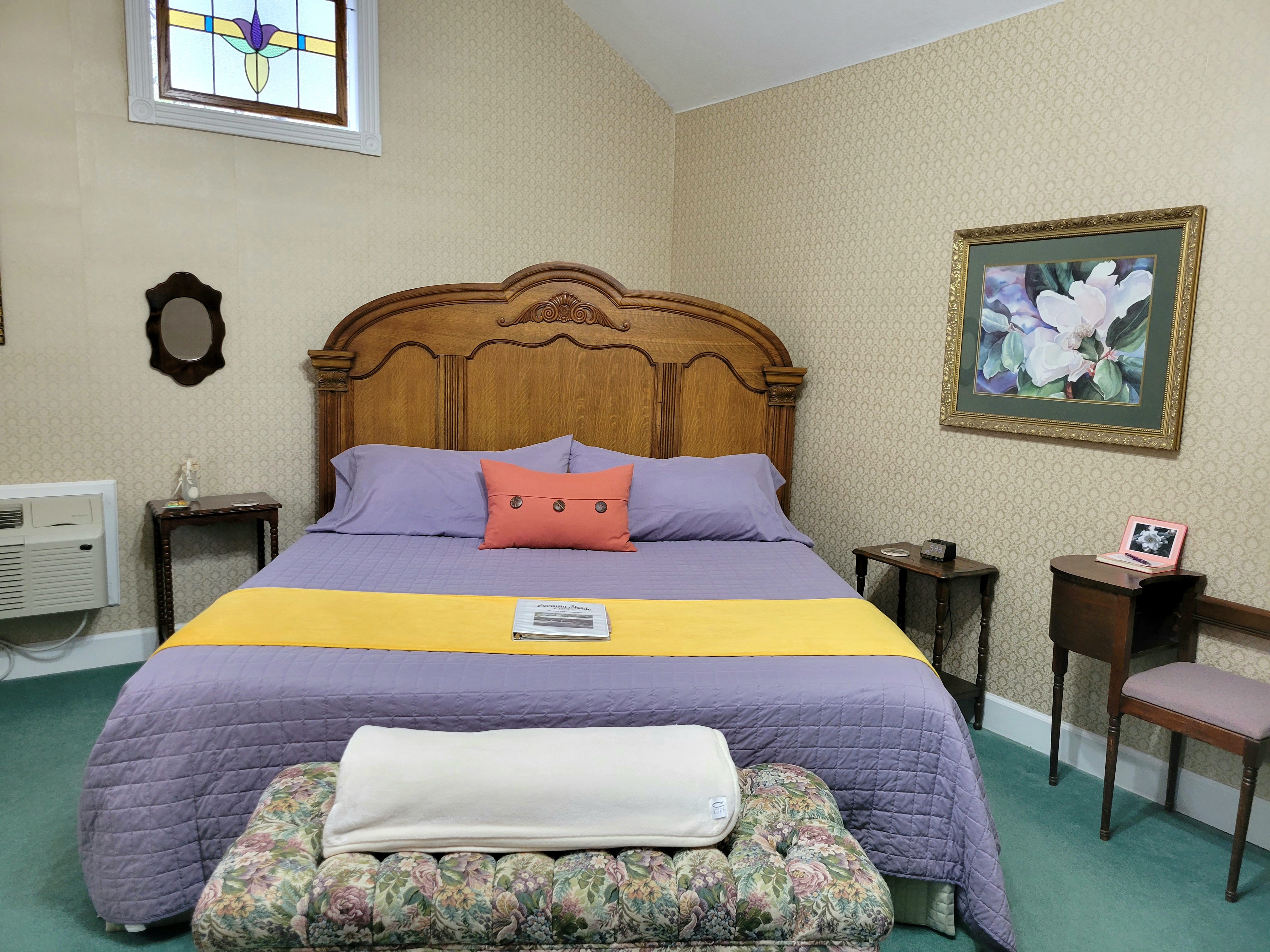 A charming suite featuring a large, ornate wooden king-size bed with purple linens and a yellow runner. The room is decorated in a vintage style with green carpeting, a framed floral painting on the wall, and a colorful stained-glass window accent above the headboard.