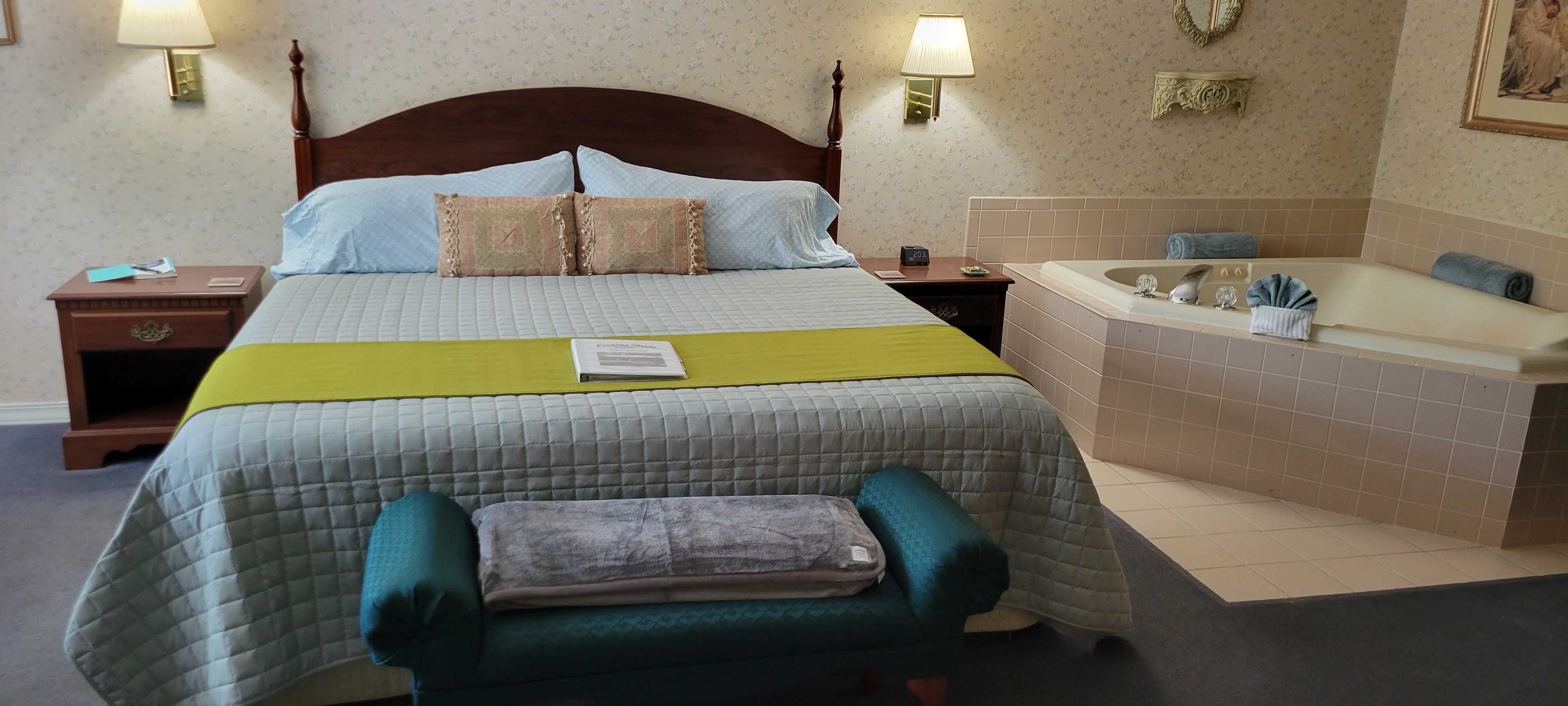 A spacious bedroom featuring a king-size bed with a green runner, flanked by wooden nightstands and wall-mounted lamps. A large, two-person jetted tub is built into the corner of the room, adjacent to the bed.