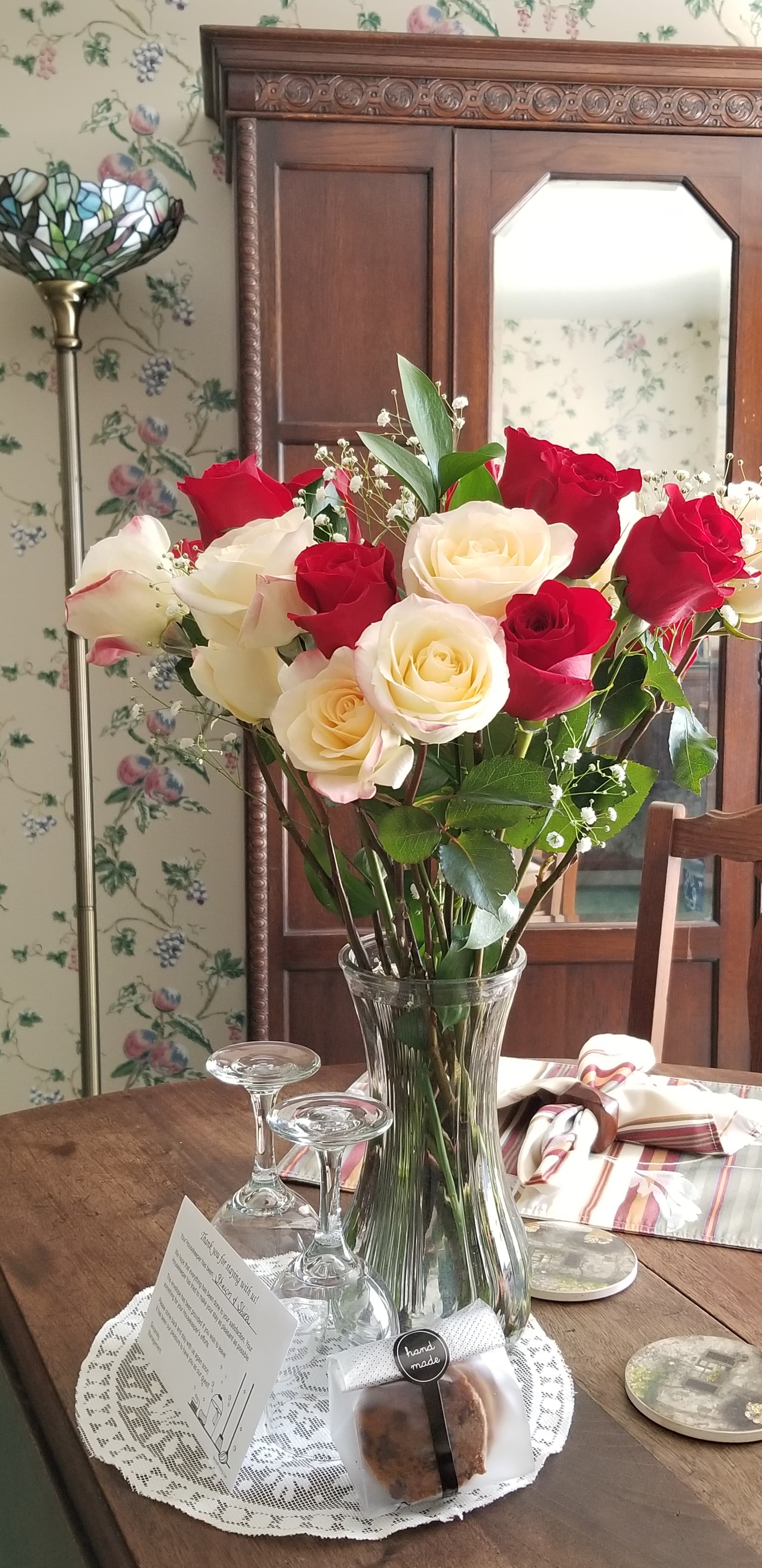 An interior close-up of a wooden dining table featuring a tall glass vase filled with fresh red and white roses. The table is set for a romantic meal with wine glasses on a lace doily and patterned napkins, positioned in front of a vintage wooden armoire and floral-patterned wallpaper.