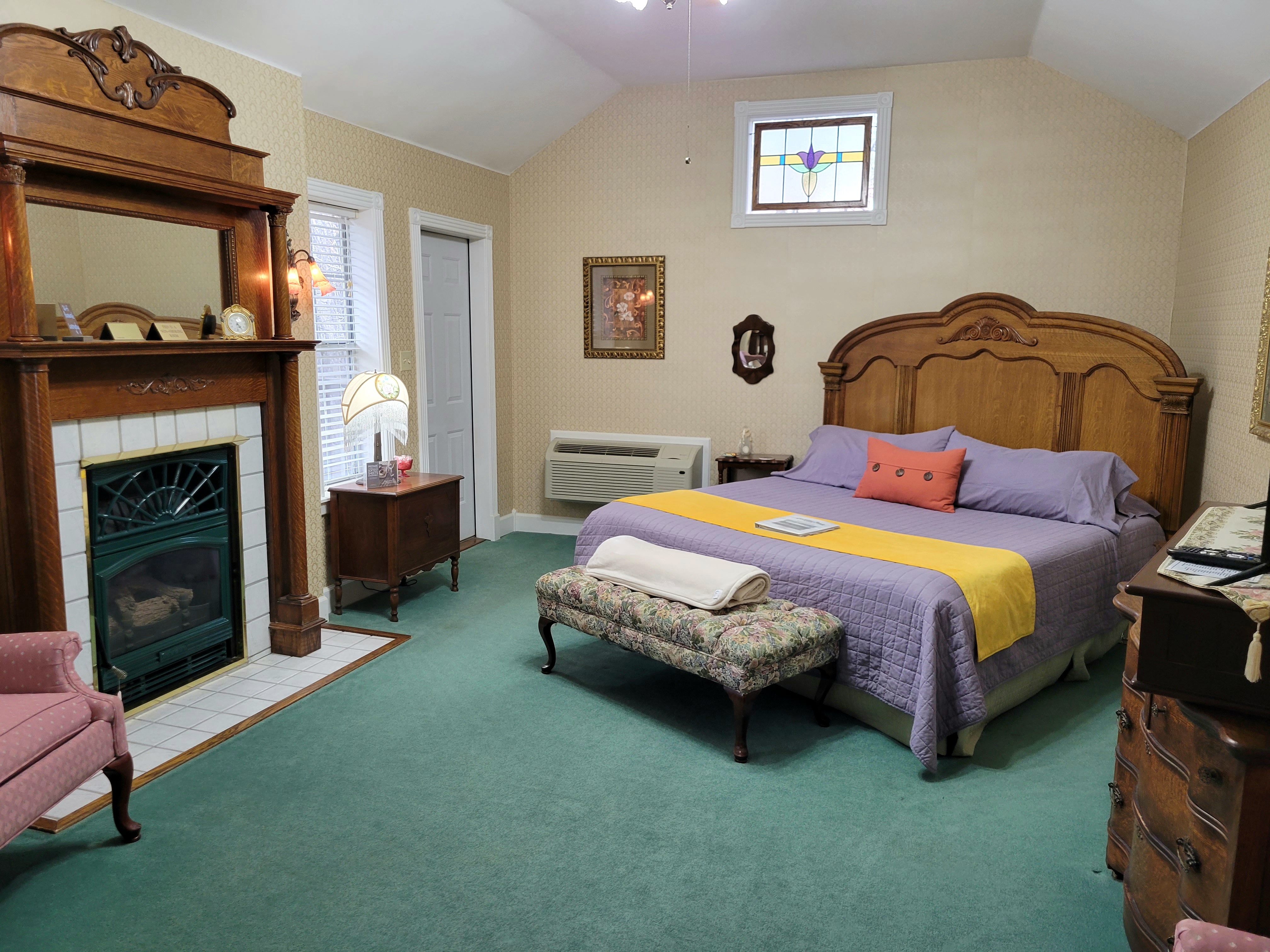 An interior view of a spacious suite featuring a large, ornate wooden king-size bed with purple linens and a yellow runner. The room includes a classic fireplace with a detailed wooden mantel, green carpeting, and a stained-glass window accent above the bed.