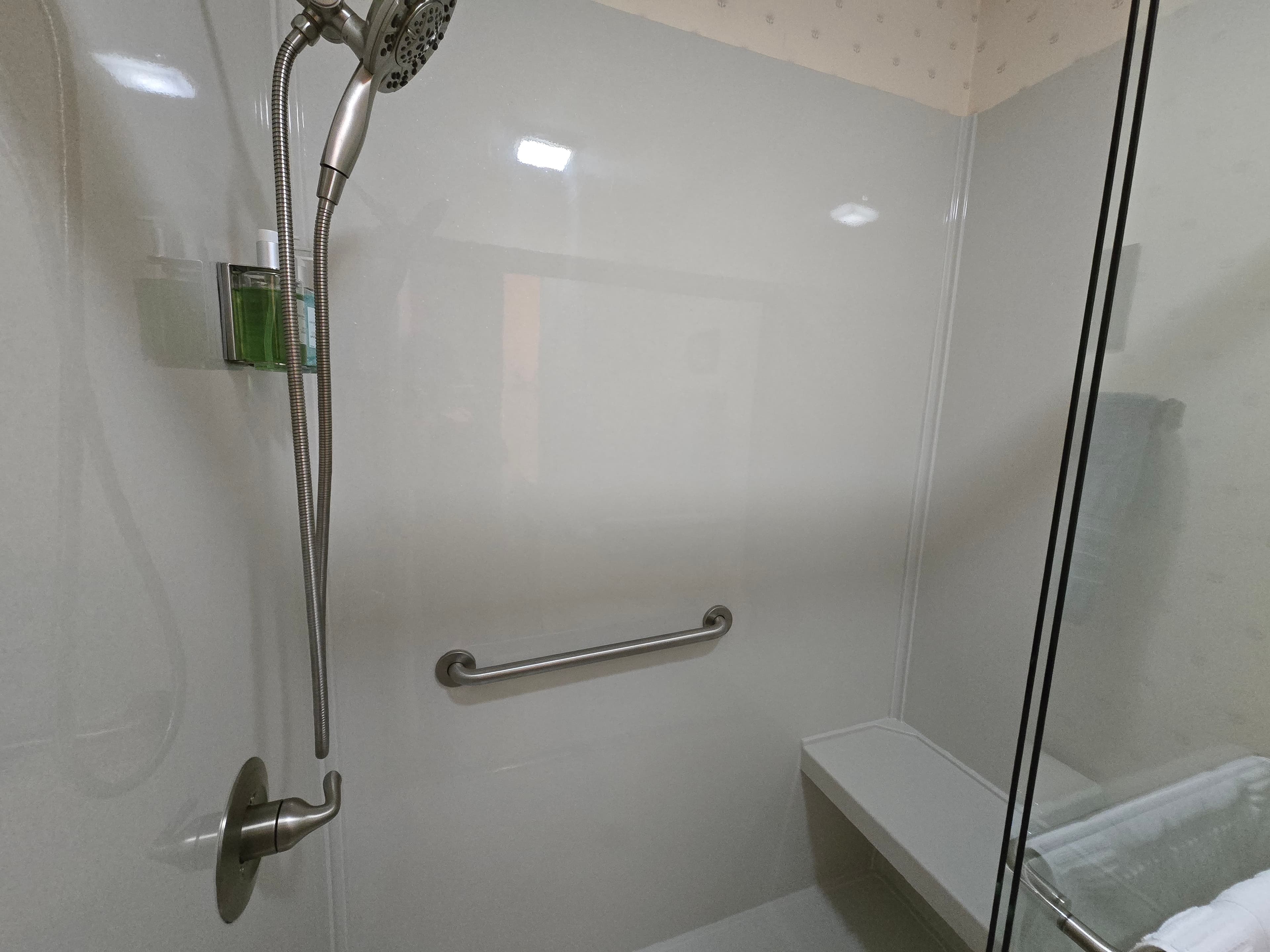 An interior view of a modern walk-in shower featuring a hand-held showerhead, a secure wall-mounted grab bar, and a built-in corner bench for comfort and accessibility.