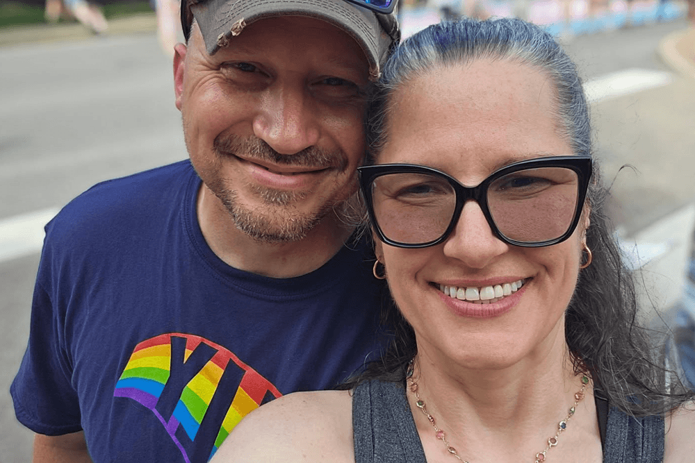 A smiling couple poses for a selfie outdoors, with a vibrant rainbow design on the man's shirt.