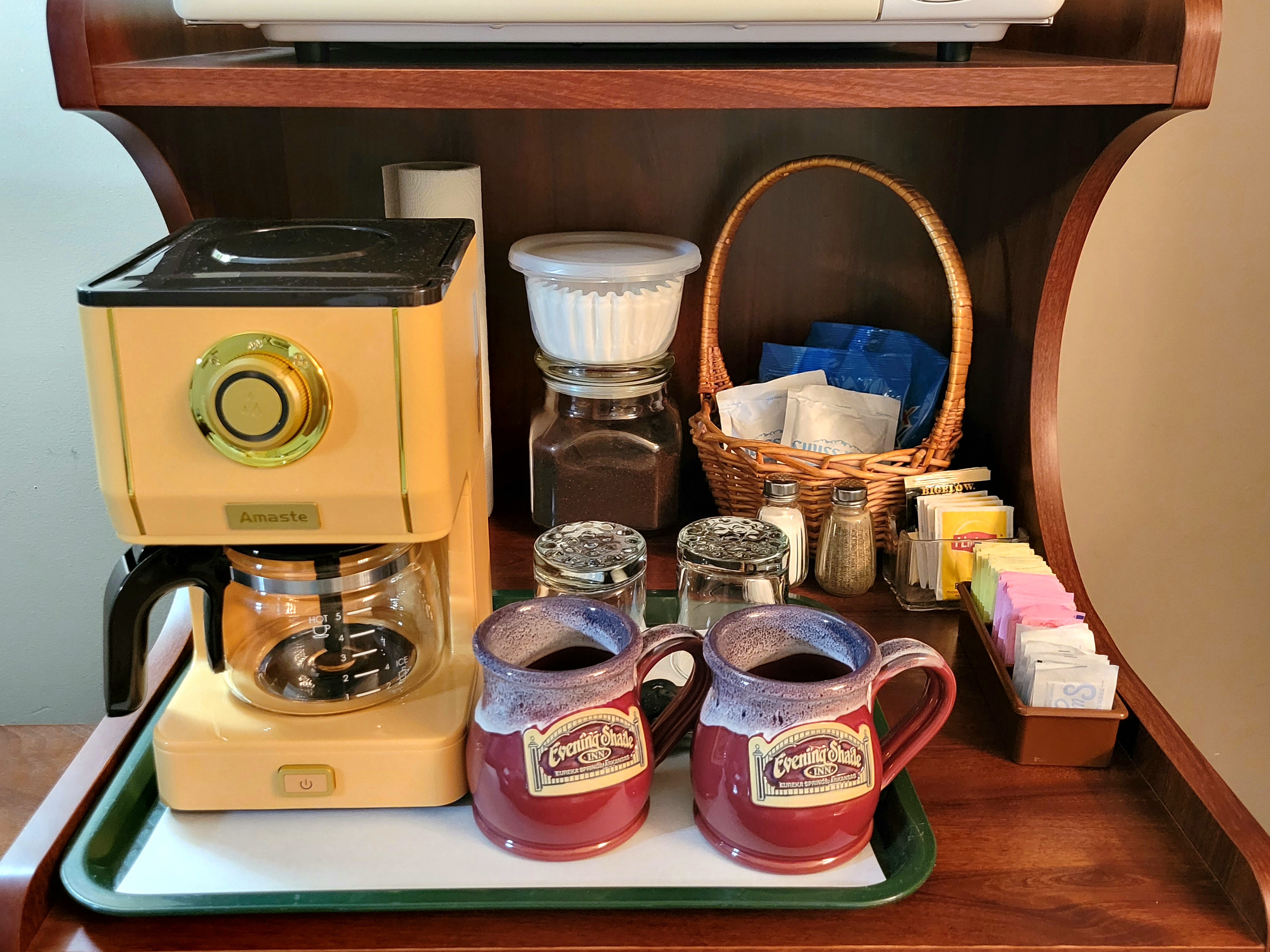 A wooden refreshment station featuring a vintage-style yellow coffee maker on a green tray with two red ceramic mugs. The station is also stocked with a basket of supplies, a glass jar of coffee grounds, and a colorful assortment of tea and sugar packets.