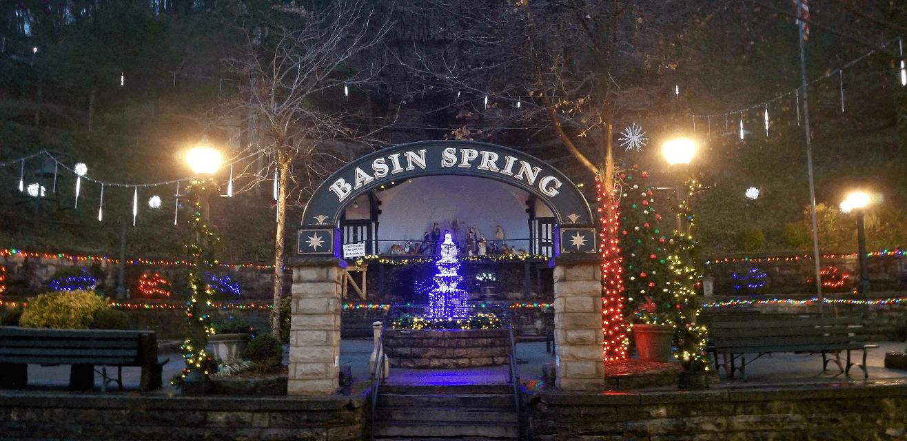 Festive scene at night in Basin Spring Park; features a glowing blue Christmas tree, colorful lights, and decorated trees under an archway, creating a joyful ambiance.