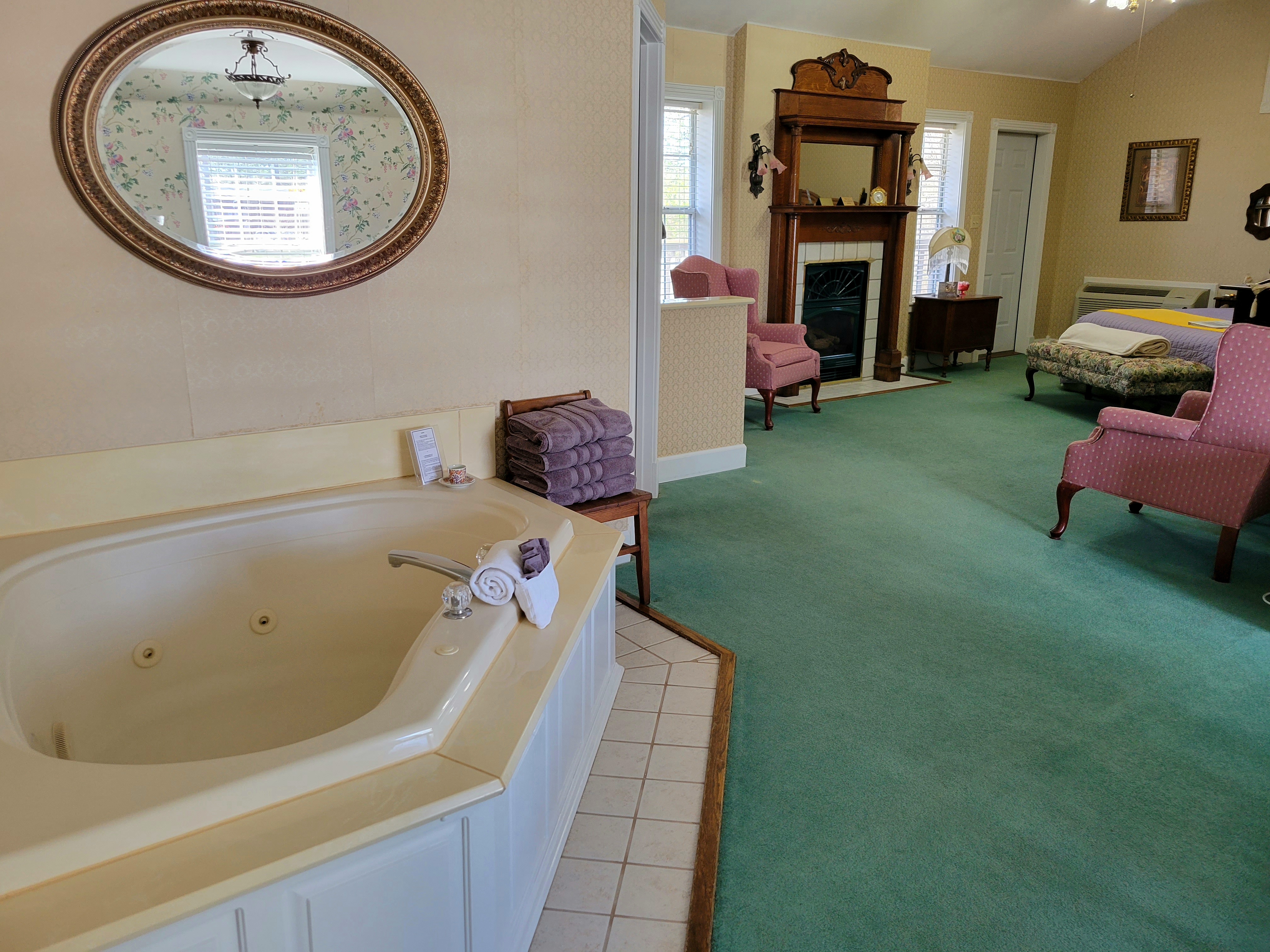 A suite showing an oval jetted tub in the foreground. The open-plan room features green carpeting, two pink wingback chairs, a classic fireplace with a detailed wooden mantel, and a king-size bed in the background.
