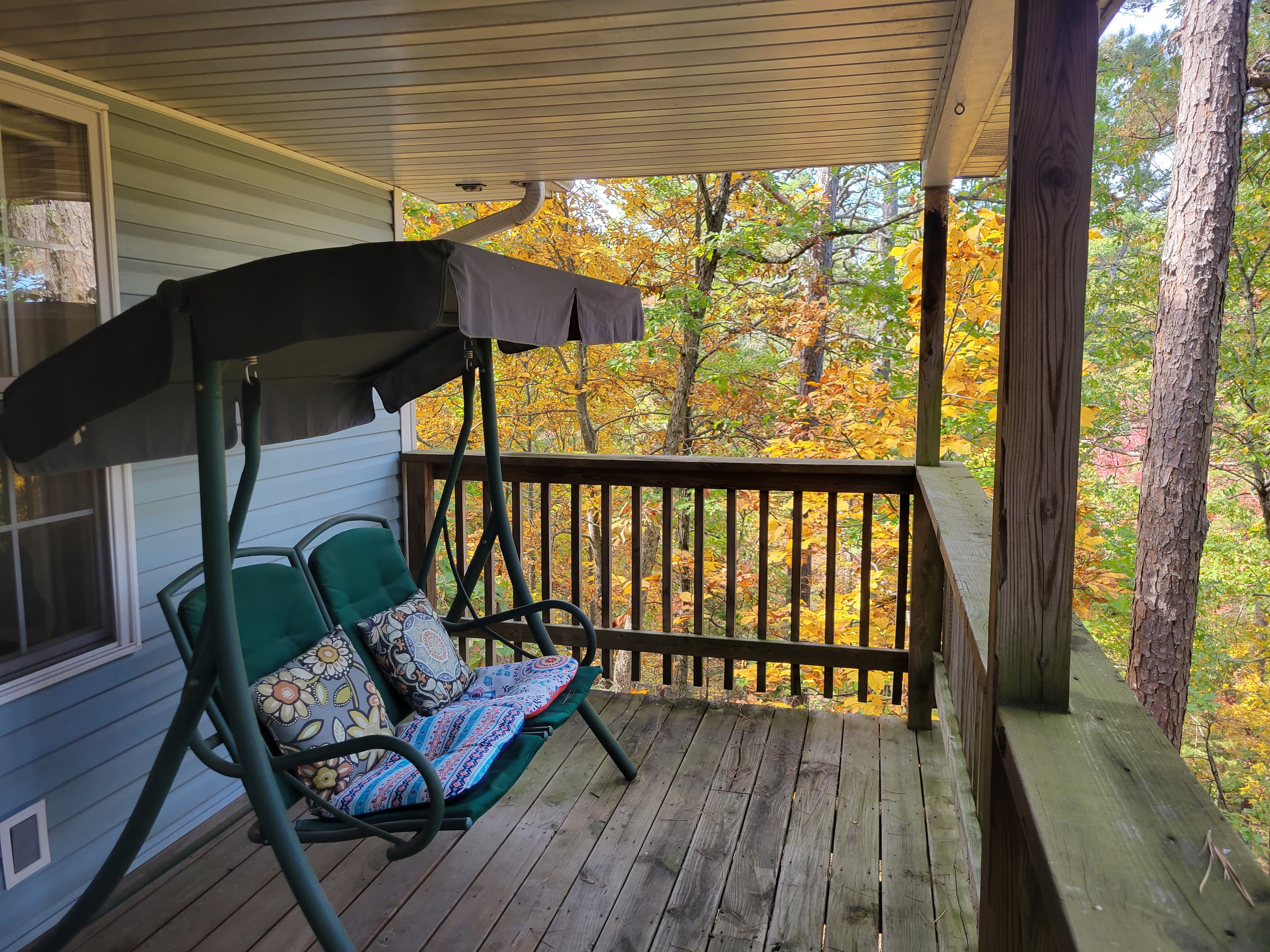 A private, covered wooden deck featuring a green cushioned swing with decorative pillows. The deck overlooks a lush forest filled with vibrant autumn foliage in shades of yellow and orange.